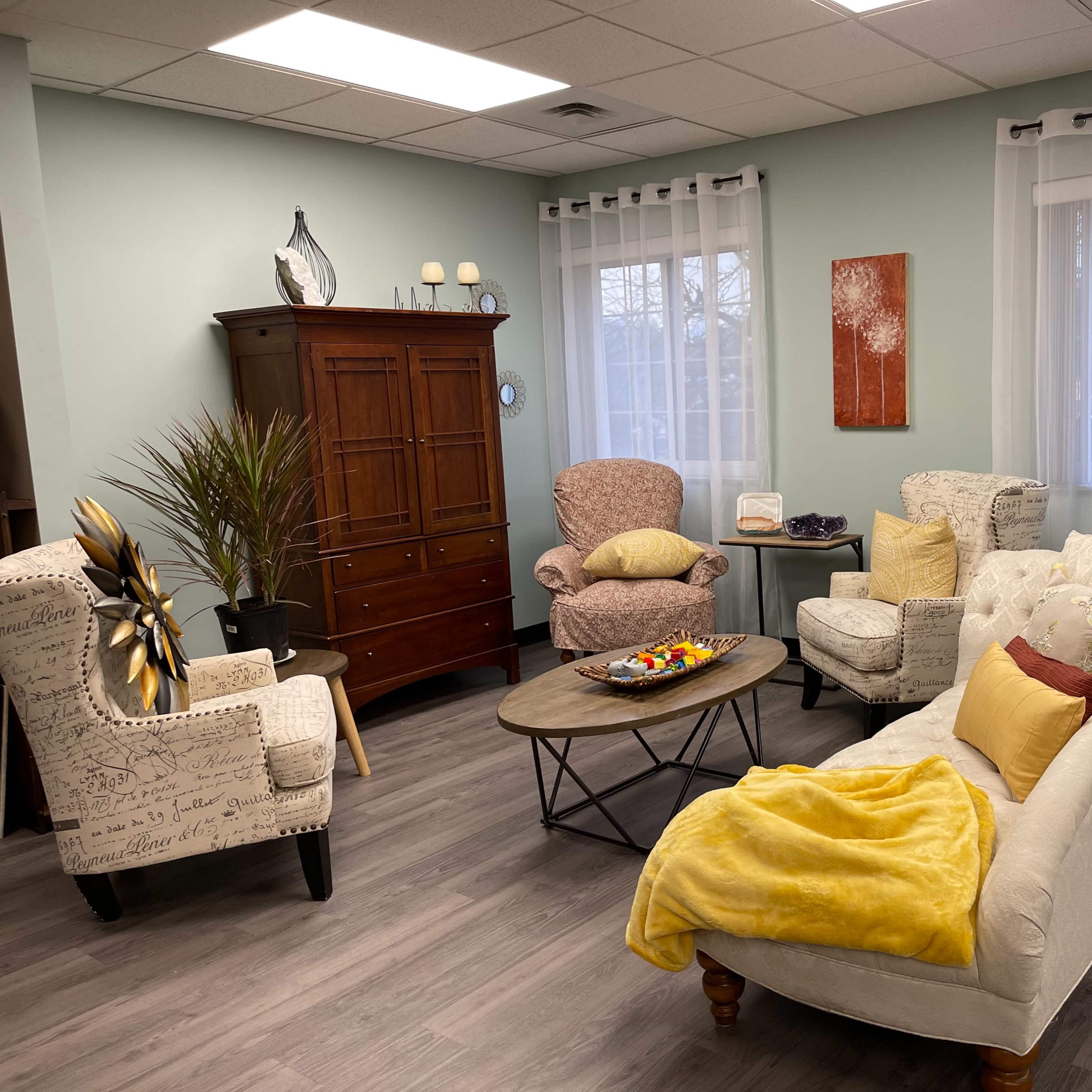 A cozy living room with light green walls, two upholstered chairs, a cream-colored sofa, a wooden cabinet, and a coffee table, all arranged around a central area with plants and decorative items.