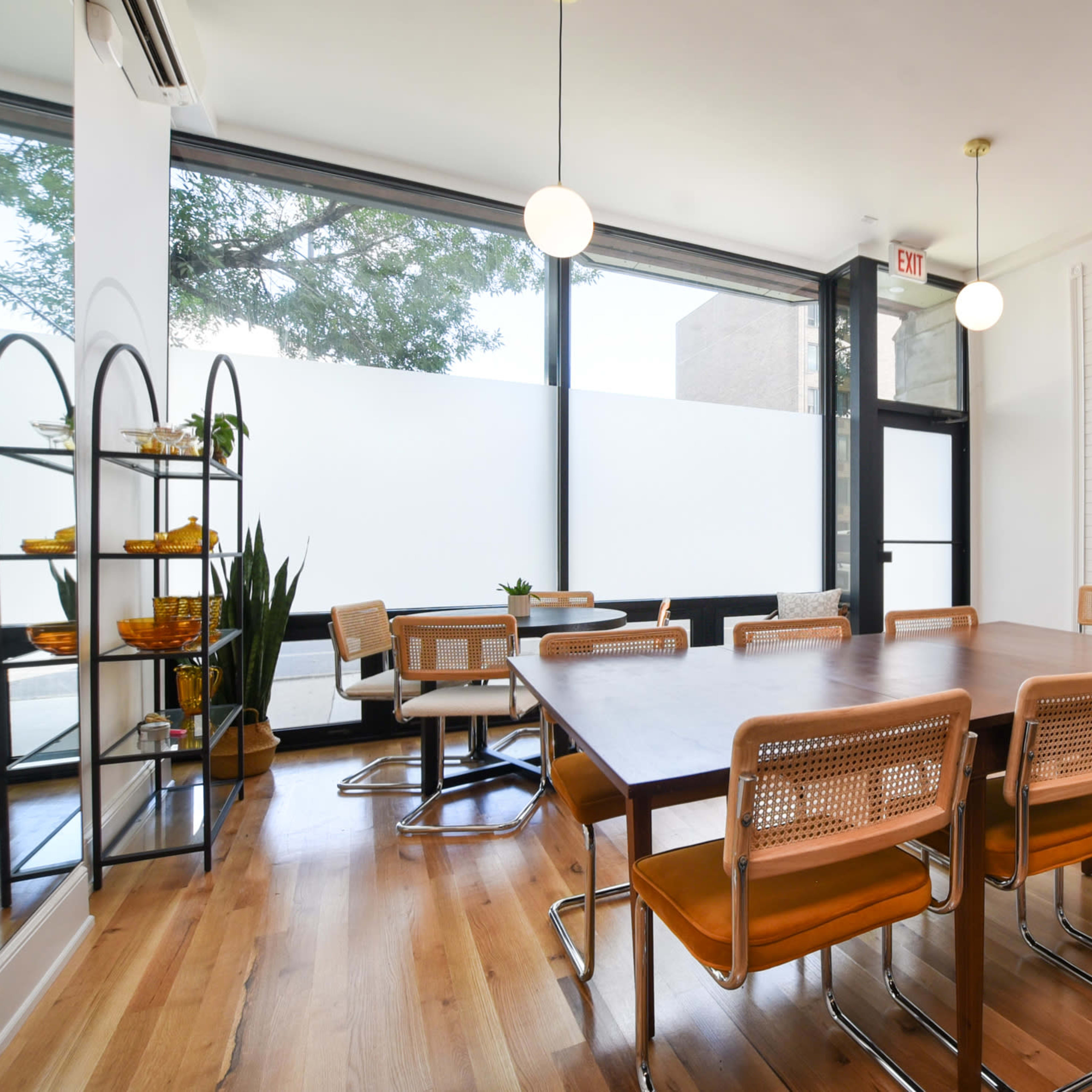 A modern dining area with a large wooden table surrounded by eight chairs, large windows letting in natural light, and a minimalist decor featuring plants and shelves.
