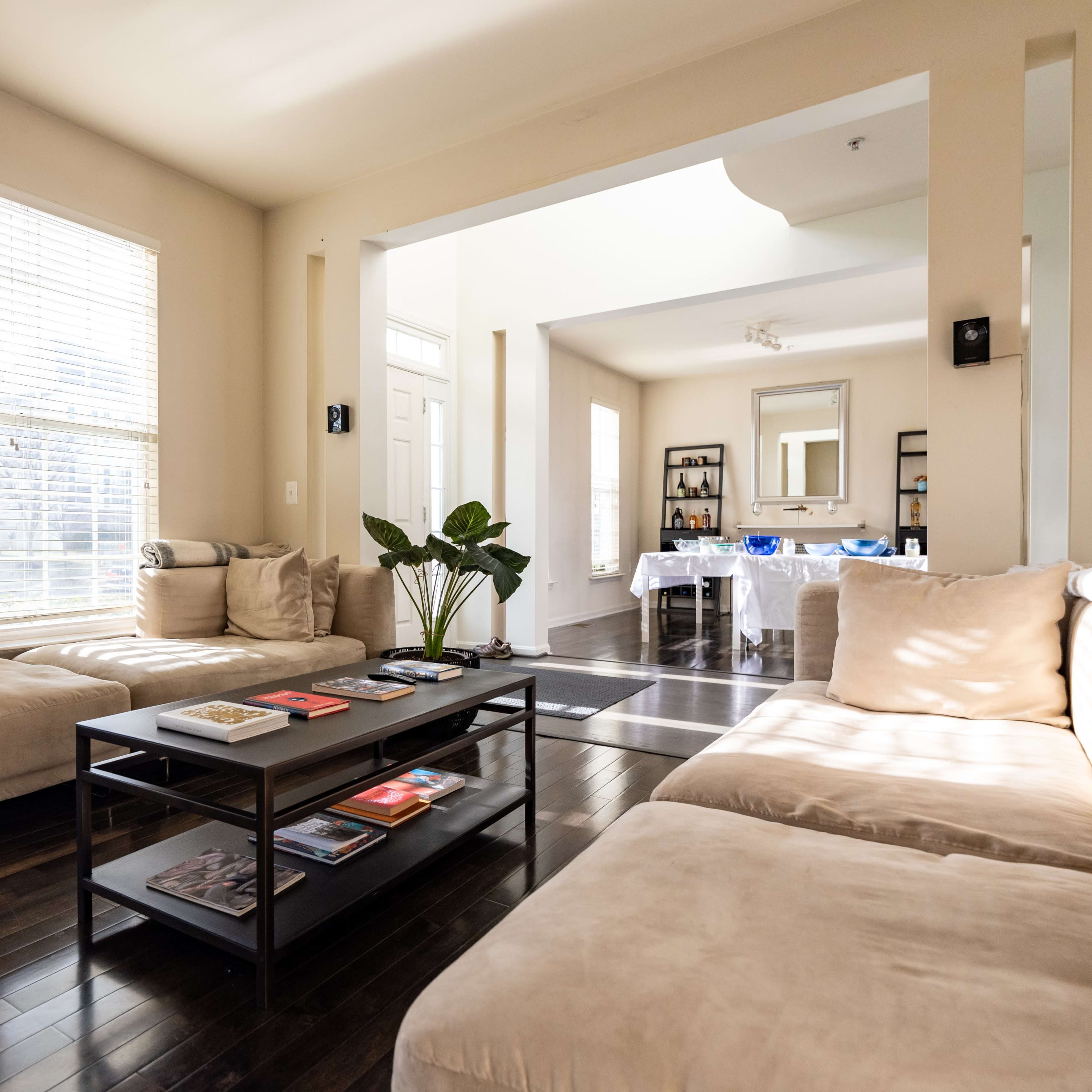 A bright living room with two sofas, a coffee table stacked with magazines, and large windows letting in natural light.