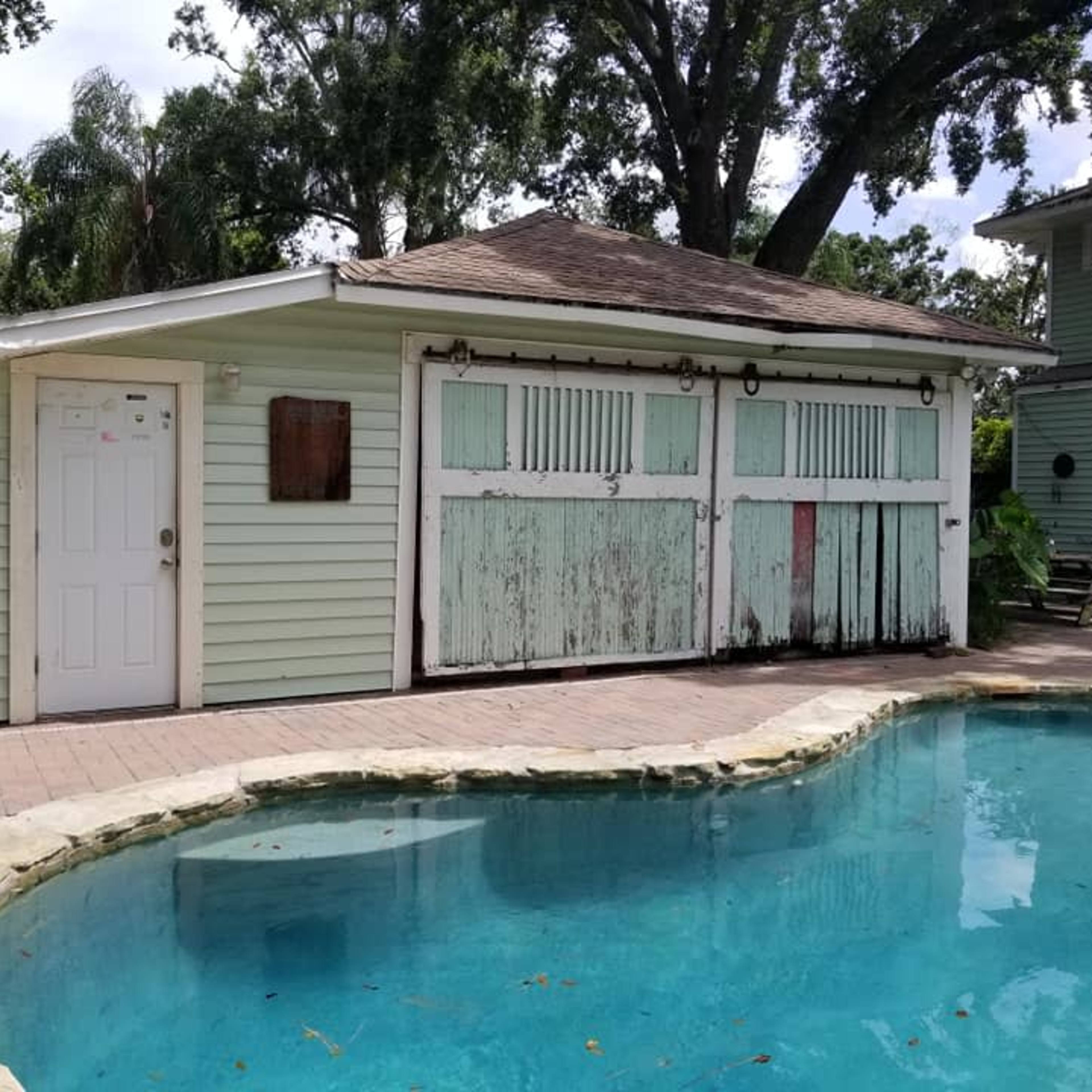 A weathered, light green shed with wooden sliding doors stands next to a blue swimming pool surrounded by patio tiles.