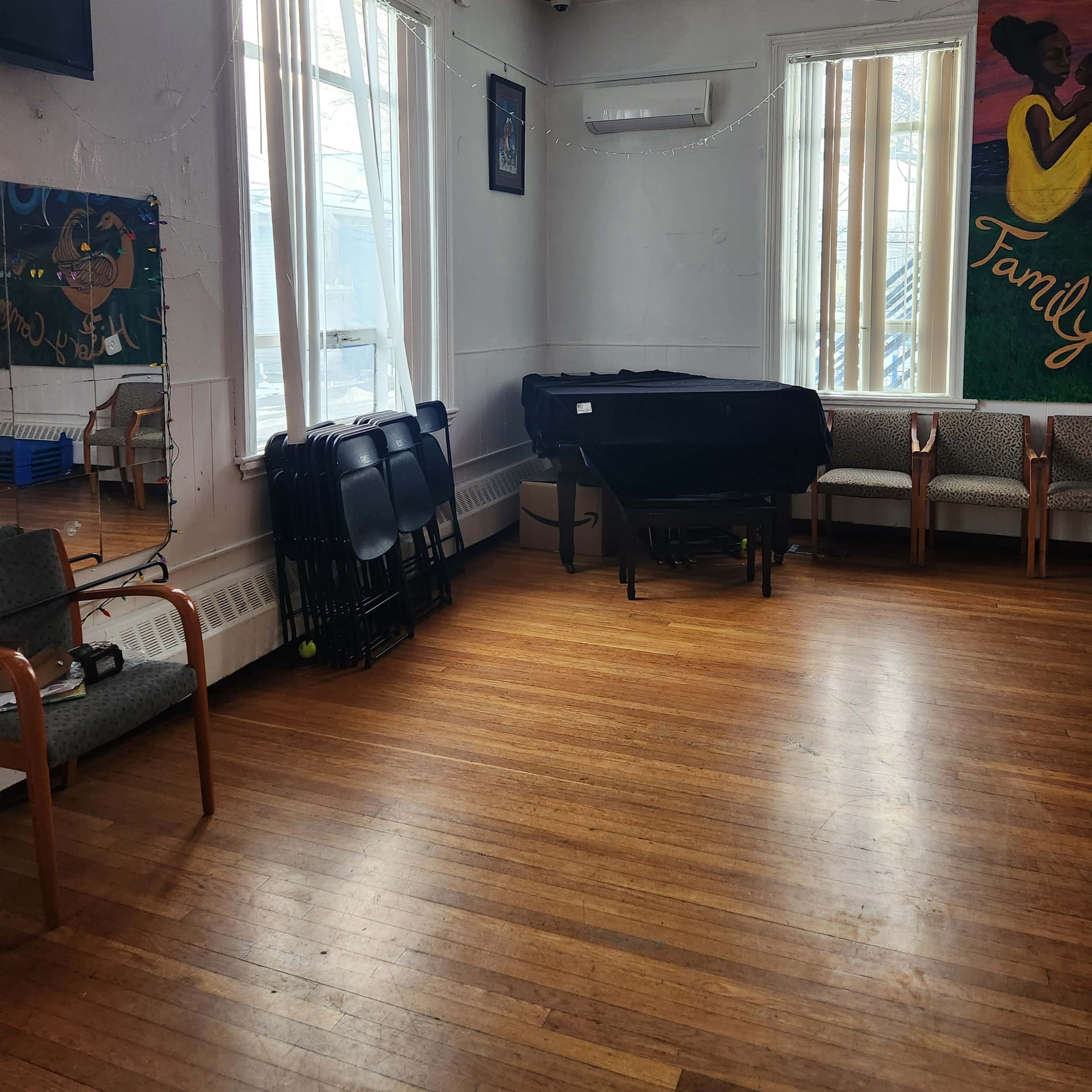 A bright room with wooden floors, a black-covered table, several stacked chairs, and two large windows allowing natural light.