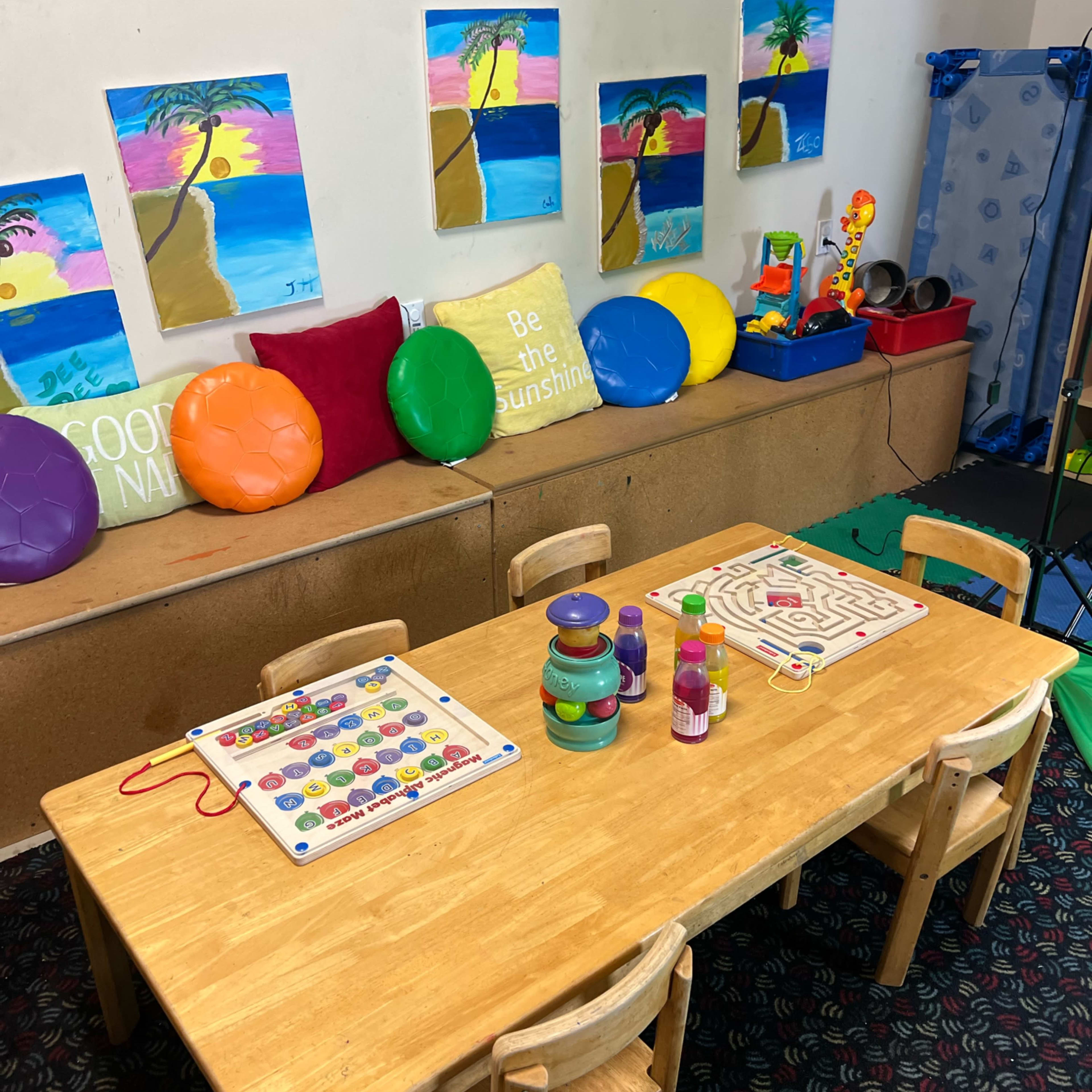 A classroom corner features a wooden table with learning games and colorful toys, surrounded by artwork depicting tropical scenes on the walls.