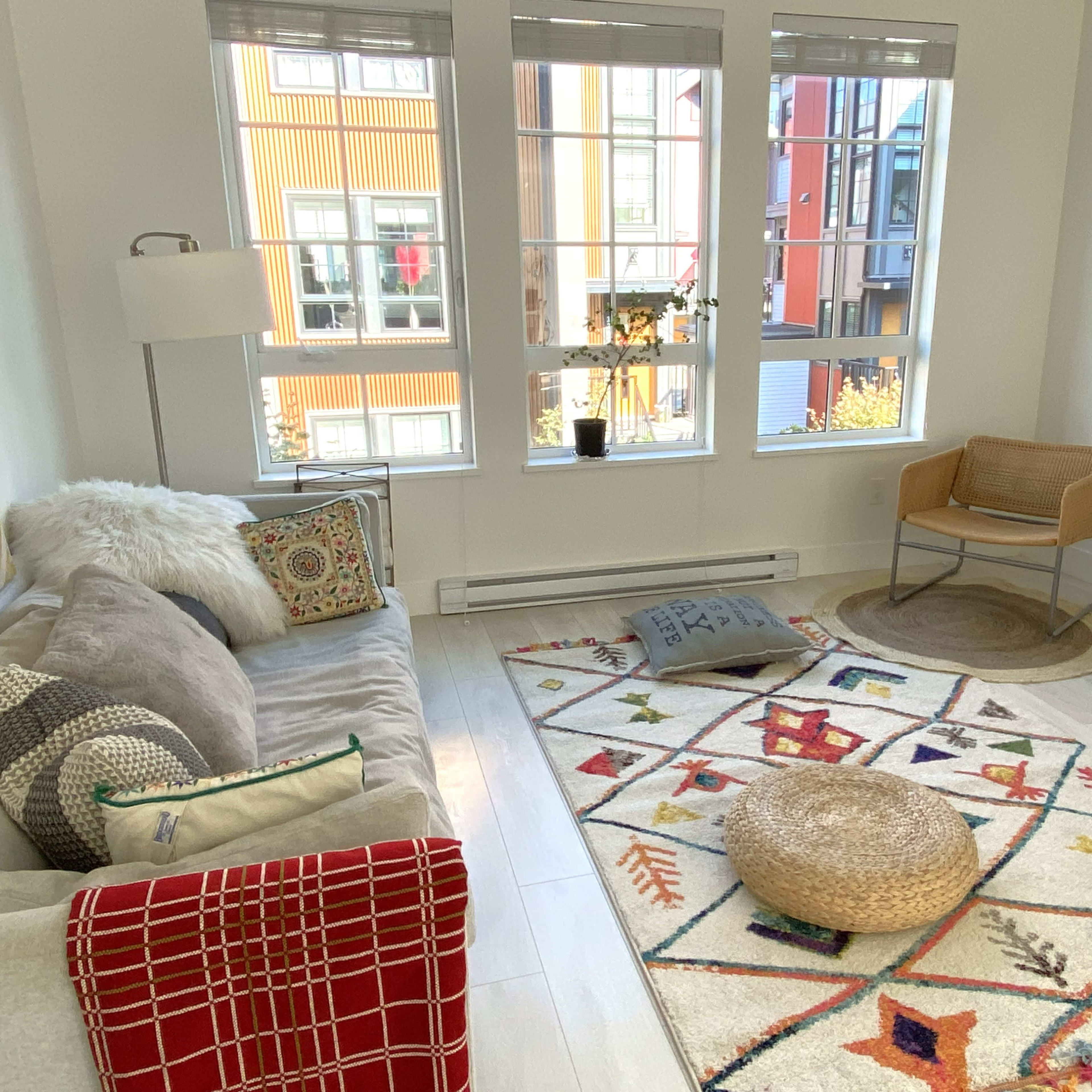 A bright living room features a light sofa, a large area rug with geometric patterns, and large windows showcasing an outside view.