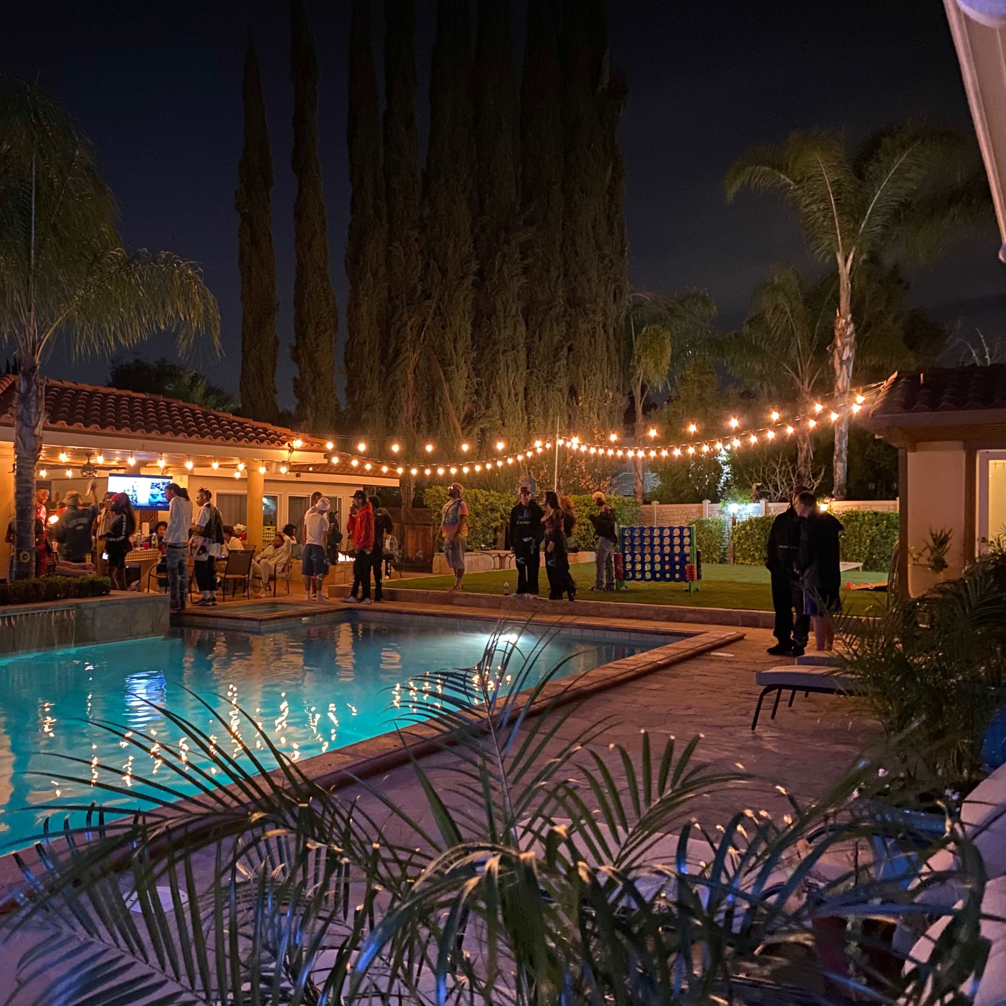A backyard party is taking place at night, featuring a pool surrounded by palm trees and string lights, with people socializing and enjoying the ambiance.