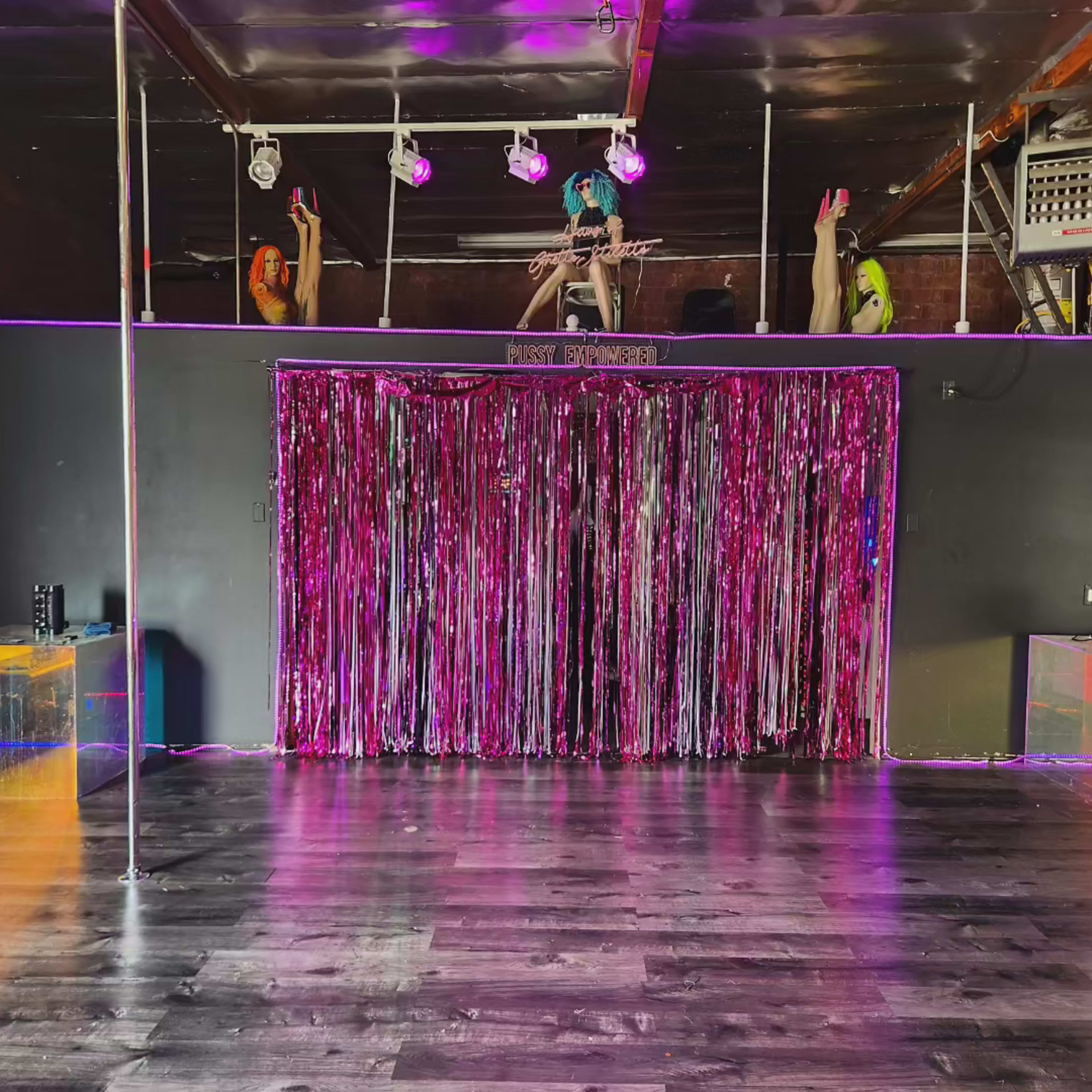 A spacious interior of a venue with a polished wooden floor, a stage adorned with colorful lighting, and a backdrop made of shiny pink streamers.