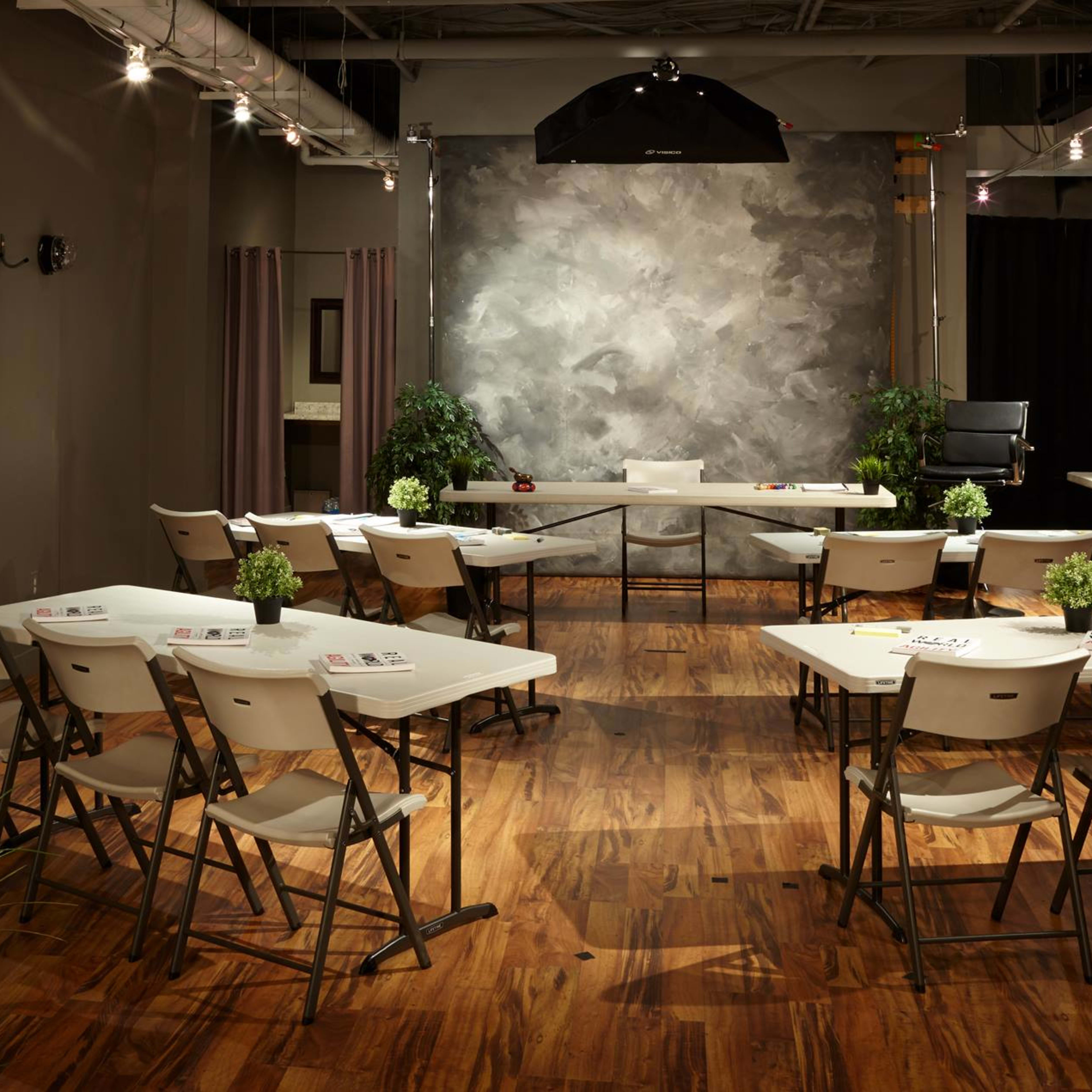 A training or meeting room is set up with rows of chairs and tables, featuring a gray backdrop and plants, illuminated by overhead lighting.