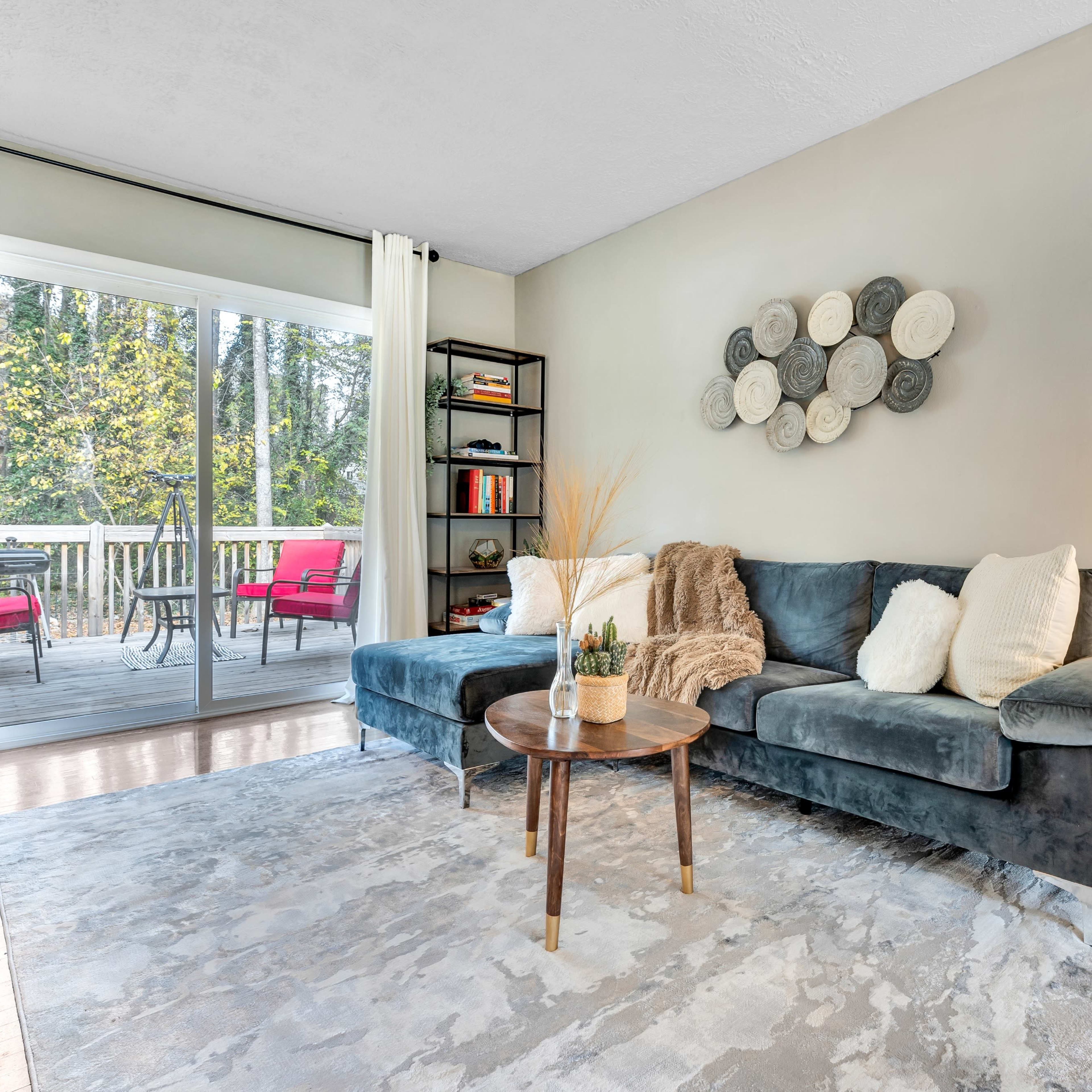 A cozy living room with a gray sectional sofa, a round coffee table, and a large window that provides a view of an outdoor deck with red chairs.