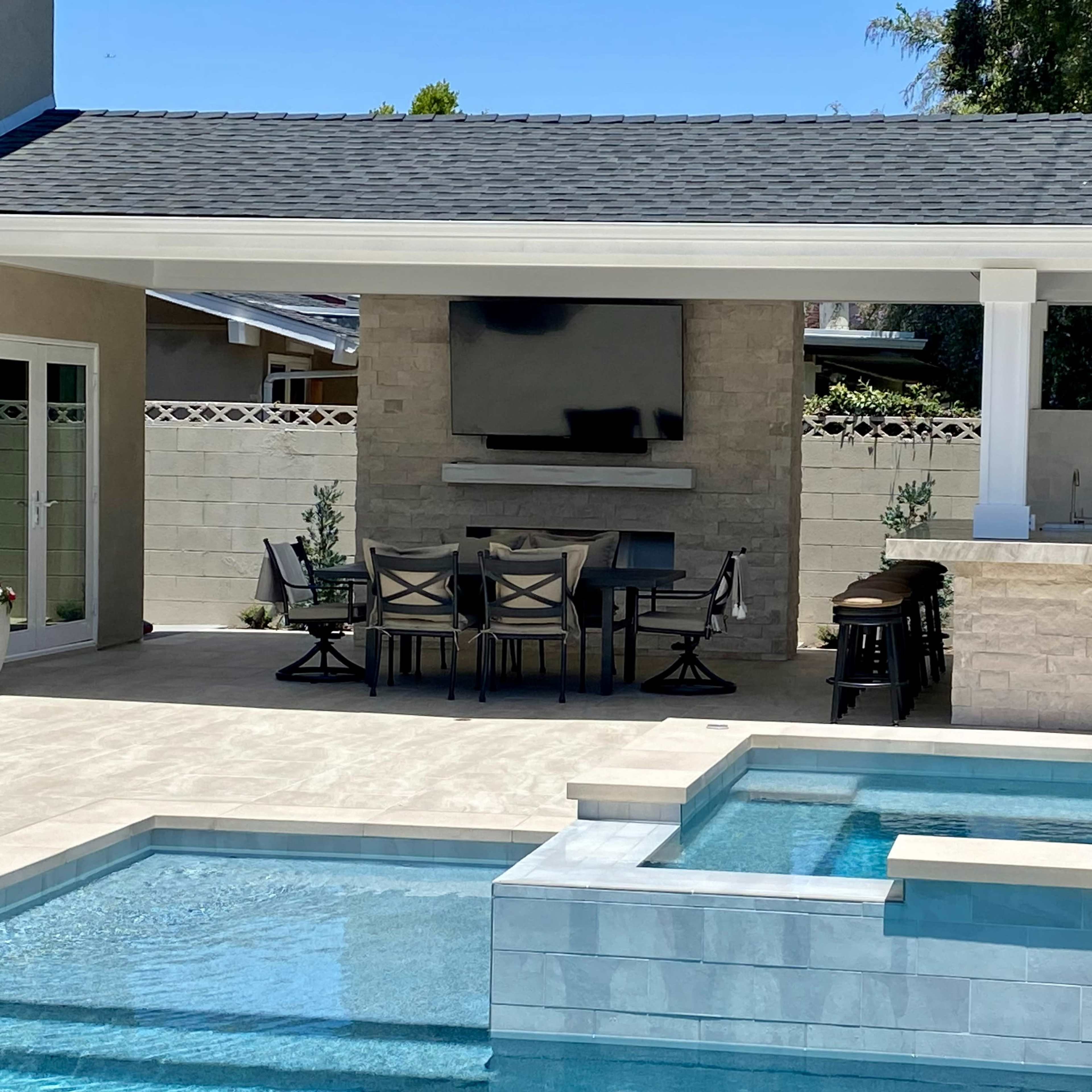 A modern outdoor patio area featuring a built-in outdoor kitchen, a dining table, and a pool with a spa.