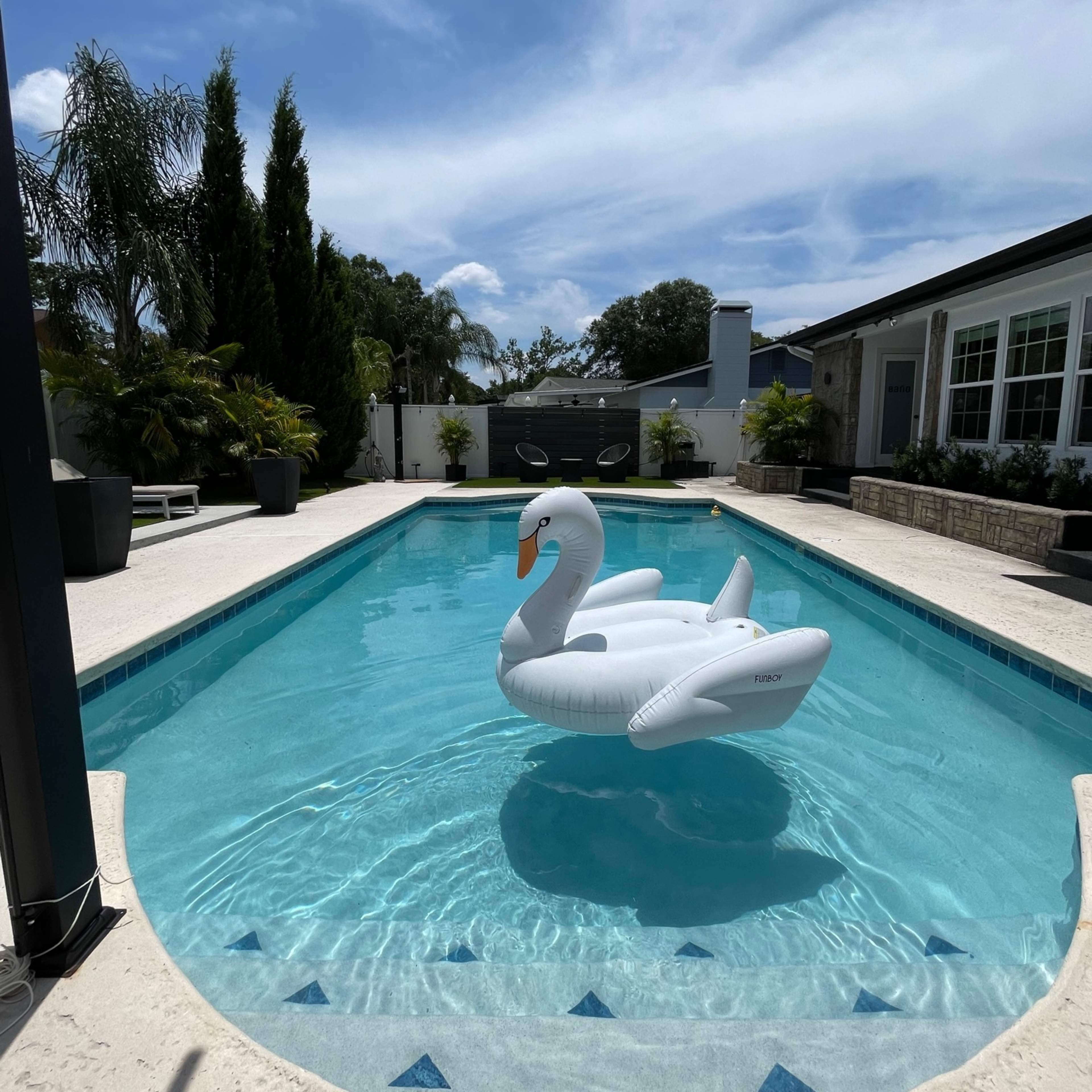 A large inflatable swan floats on a clear blue swimming pool surrounded by palm trees and a modern backyard.