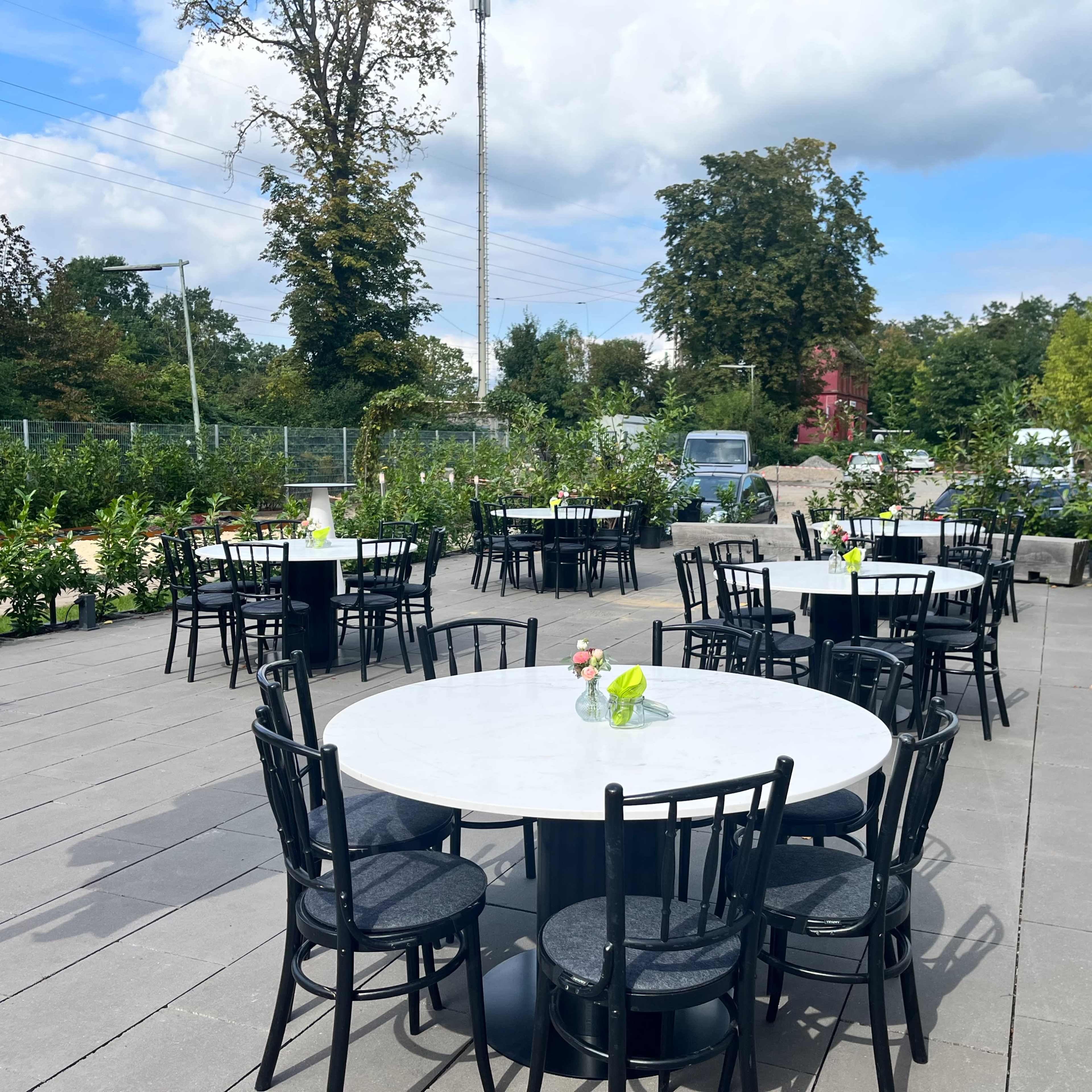 A spacious outdoor patio features several round tables with marble tops and black chairs, surrounded by greenery and under a partly cloudy sky.