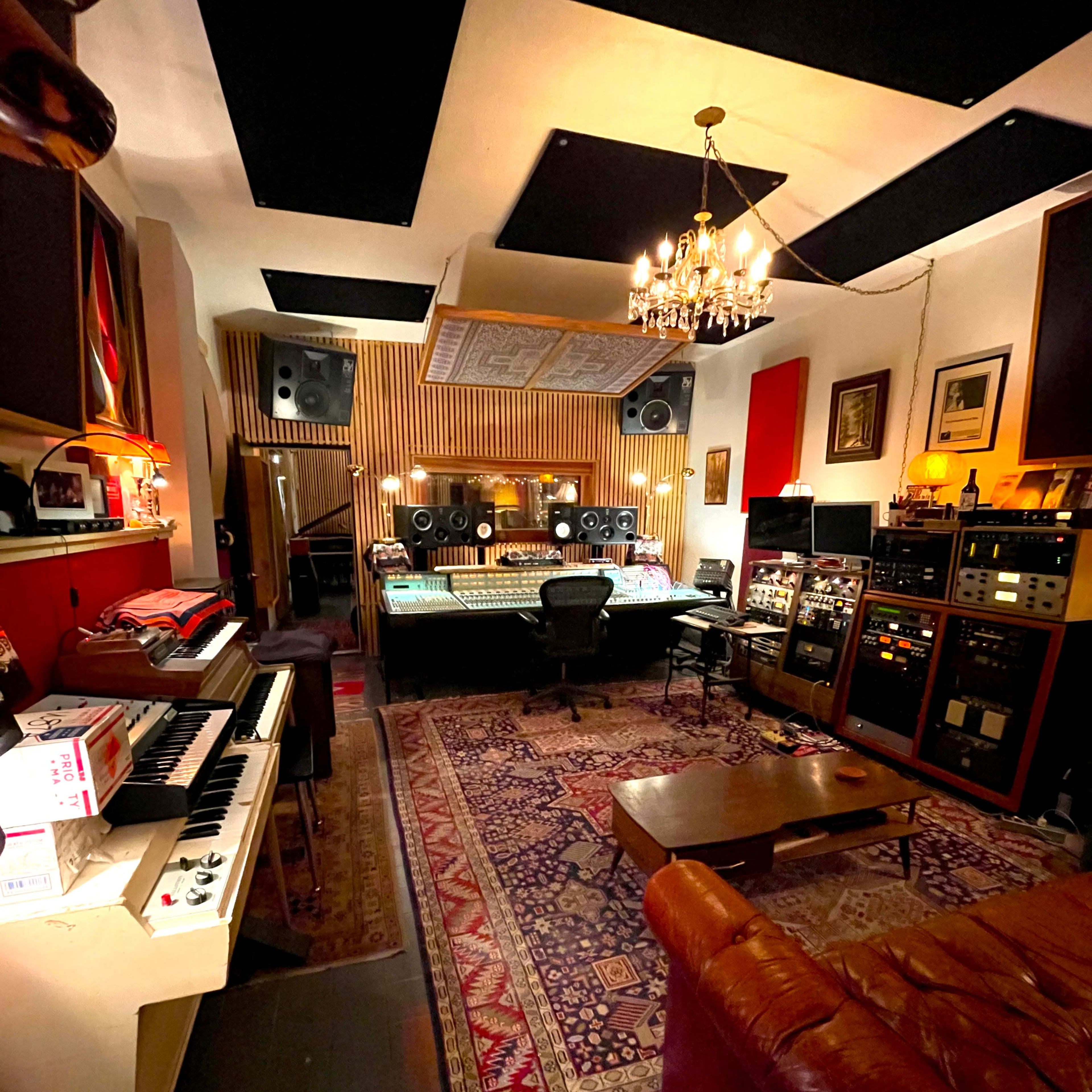 A recording studio with a large mixing console, various audio equipment, a keyboard, and a plush sofa on a patterned rug.