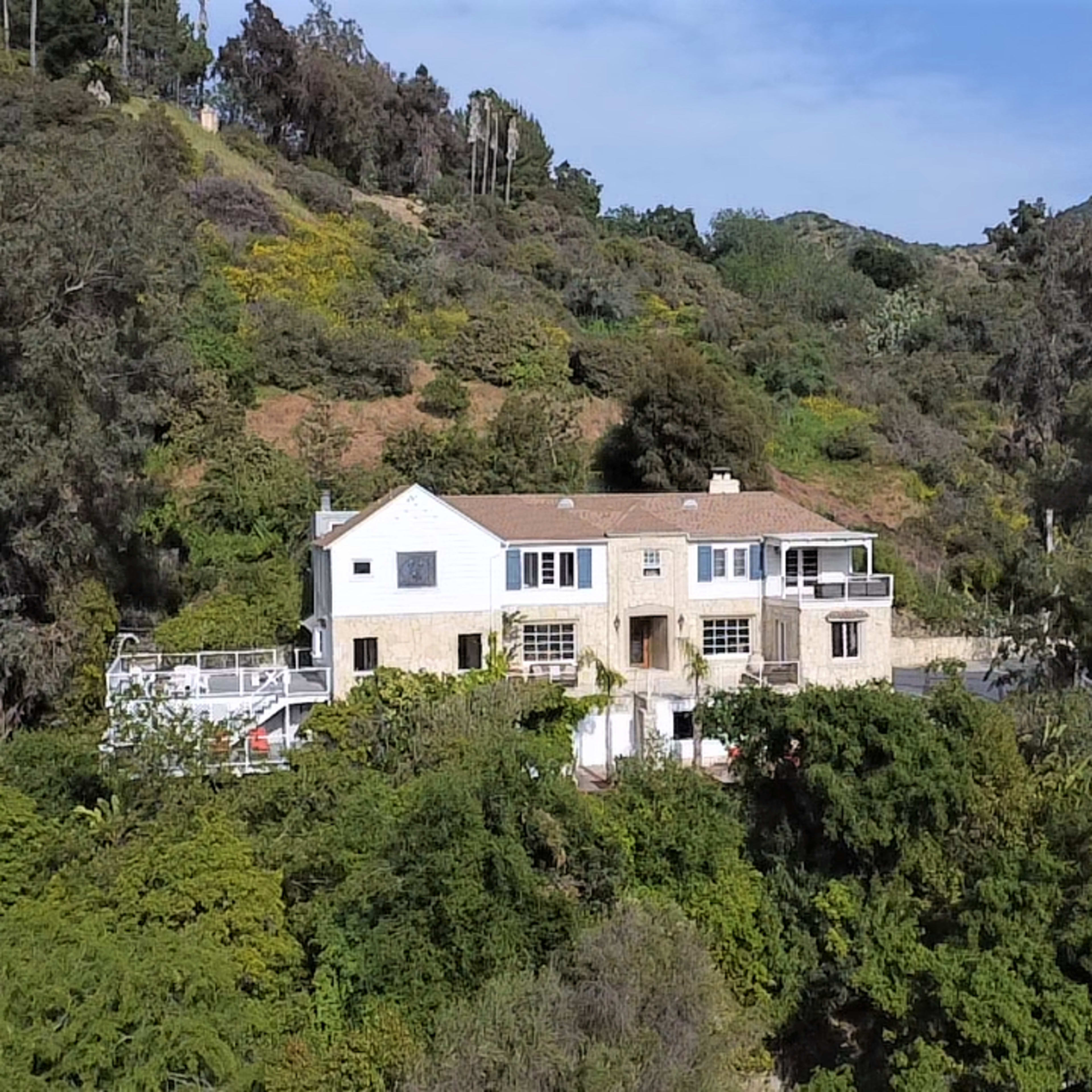 A large white house with a tan roof is surrounded by dense greenery and situated on a hillside.