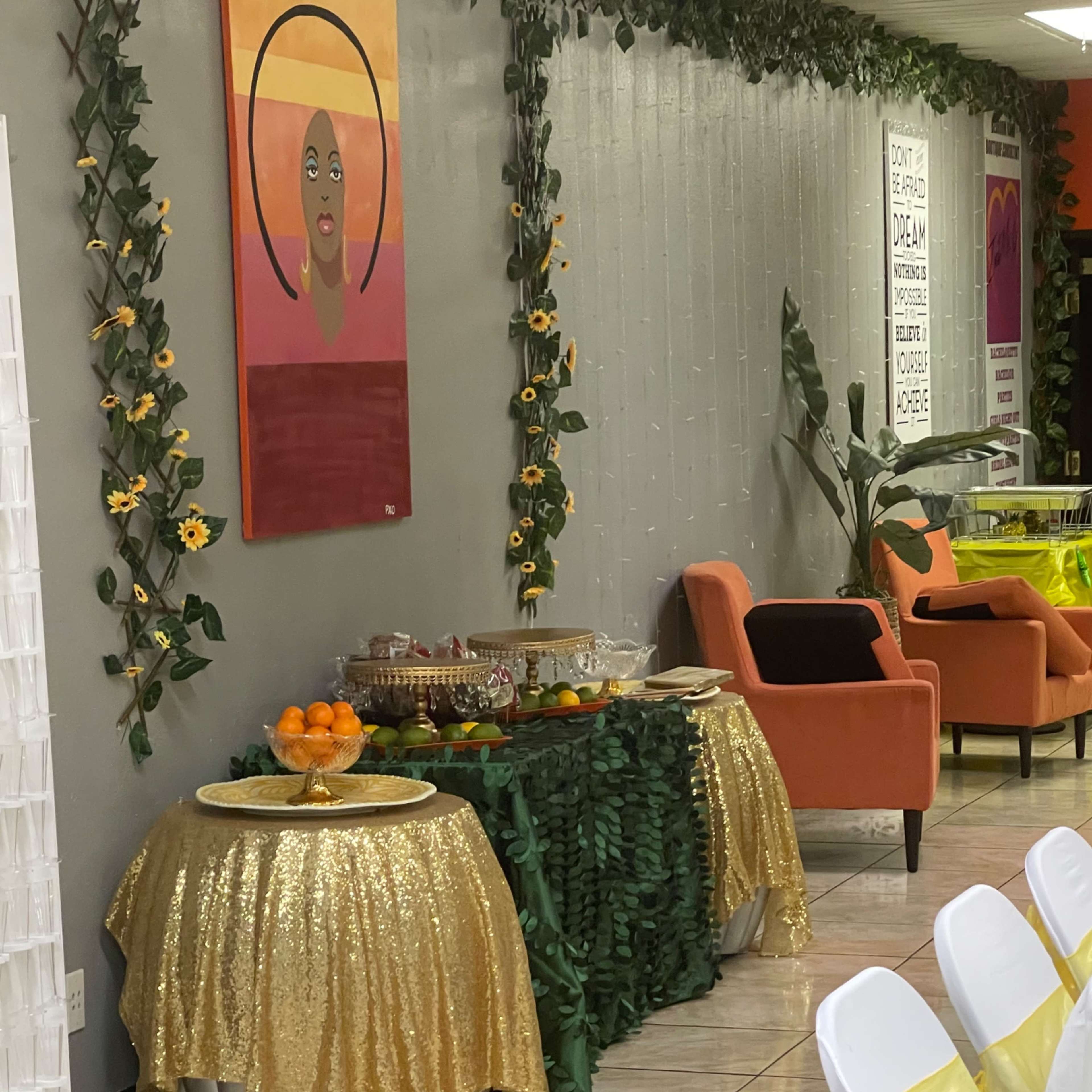 A decorated refreshment table covered with a golden tablecloth is set against a wall adorned with greenery and artwork.