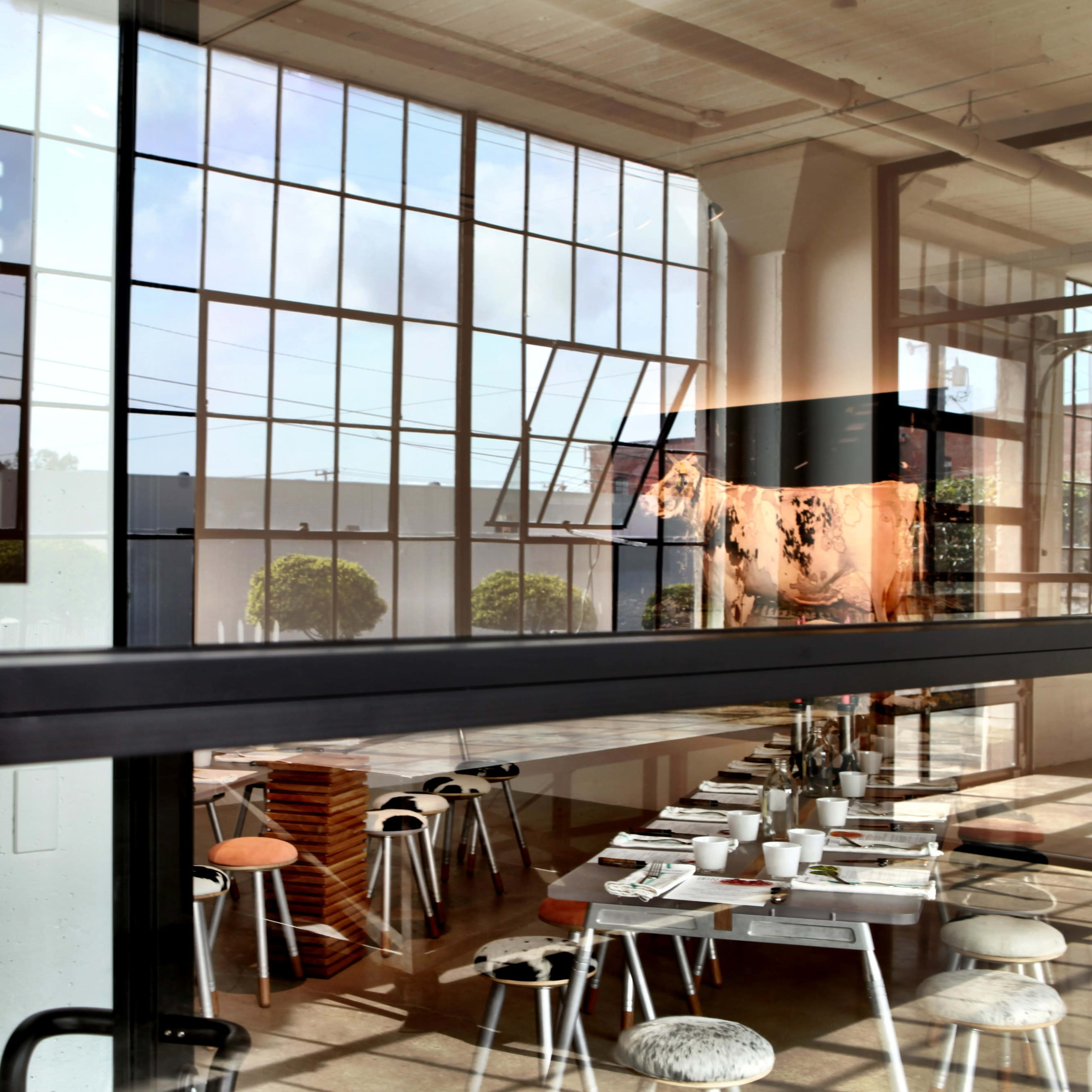 A modern art gallery with large windows and a dining area visible through the glass, featuring a long table set with plates and chairs.