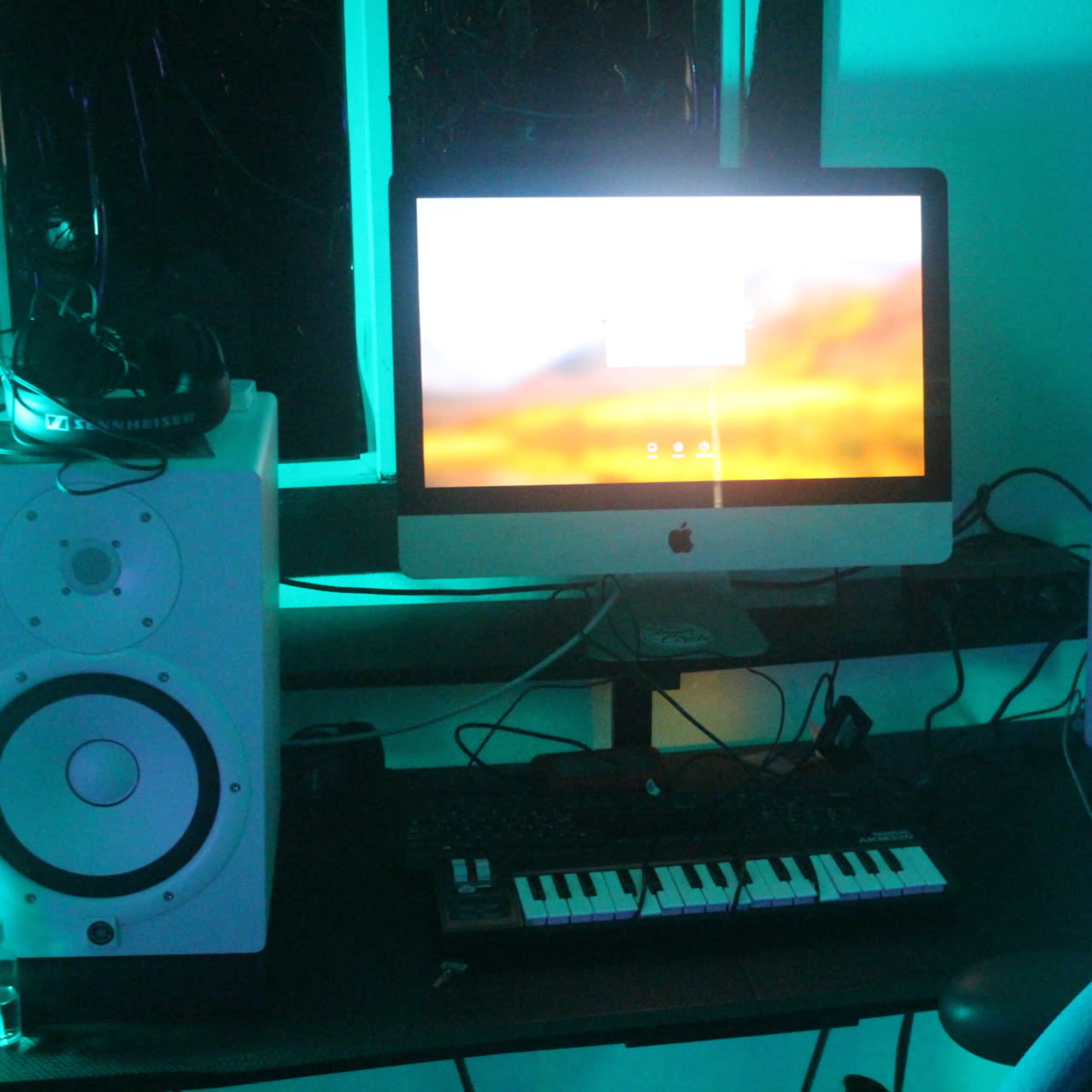 A desk holds a computer and two speakers, with a keyboard and headphones visible beside them, under blue lighting.