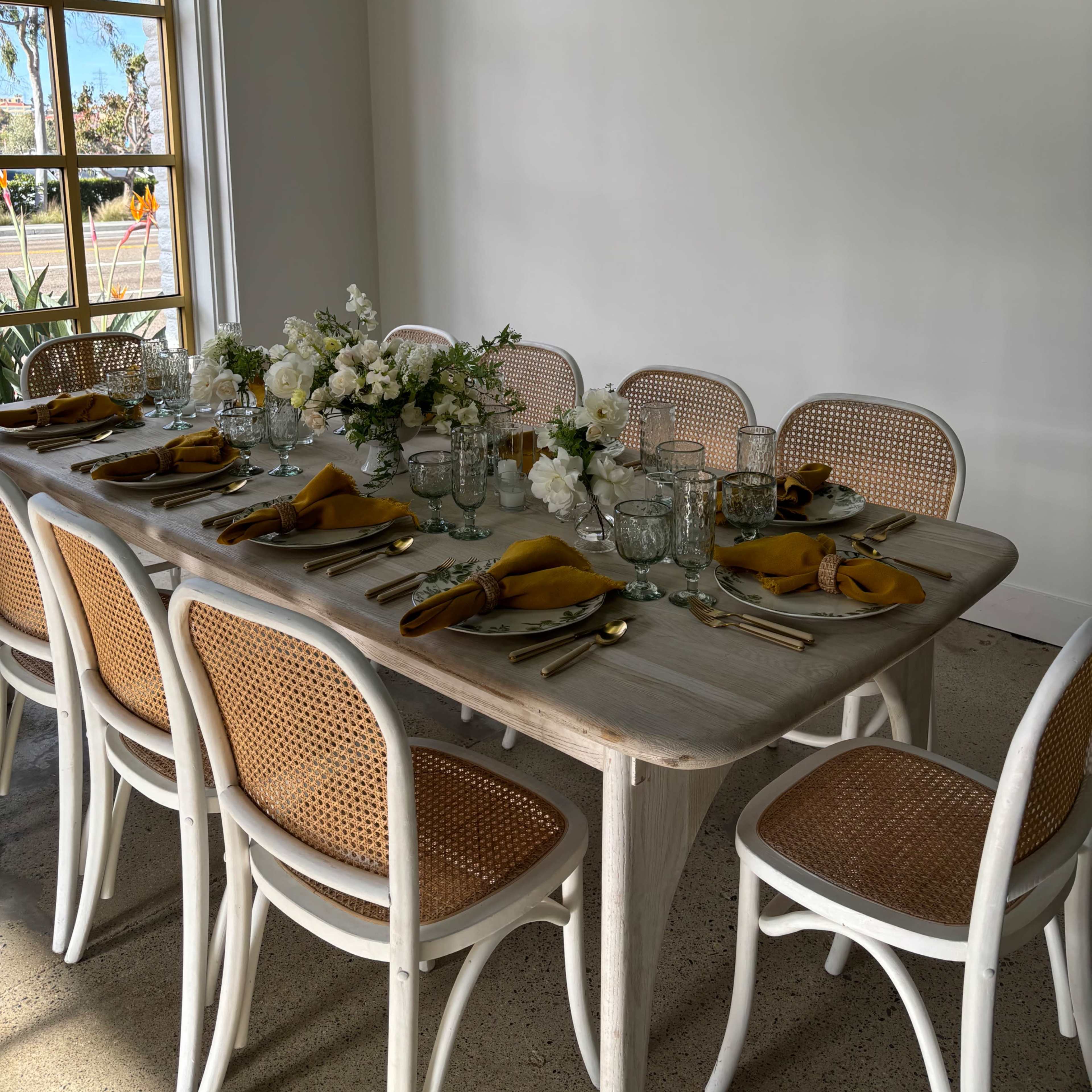 A long dining table set for a meal, adorned with glasses, plates, and a floral centerpiece, is positioned against a bright, white wall with a large window.