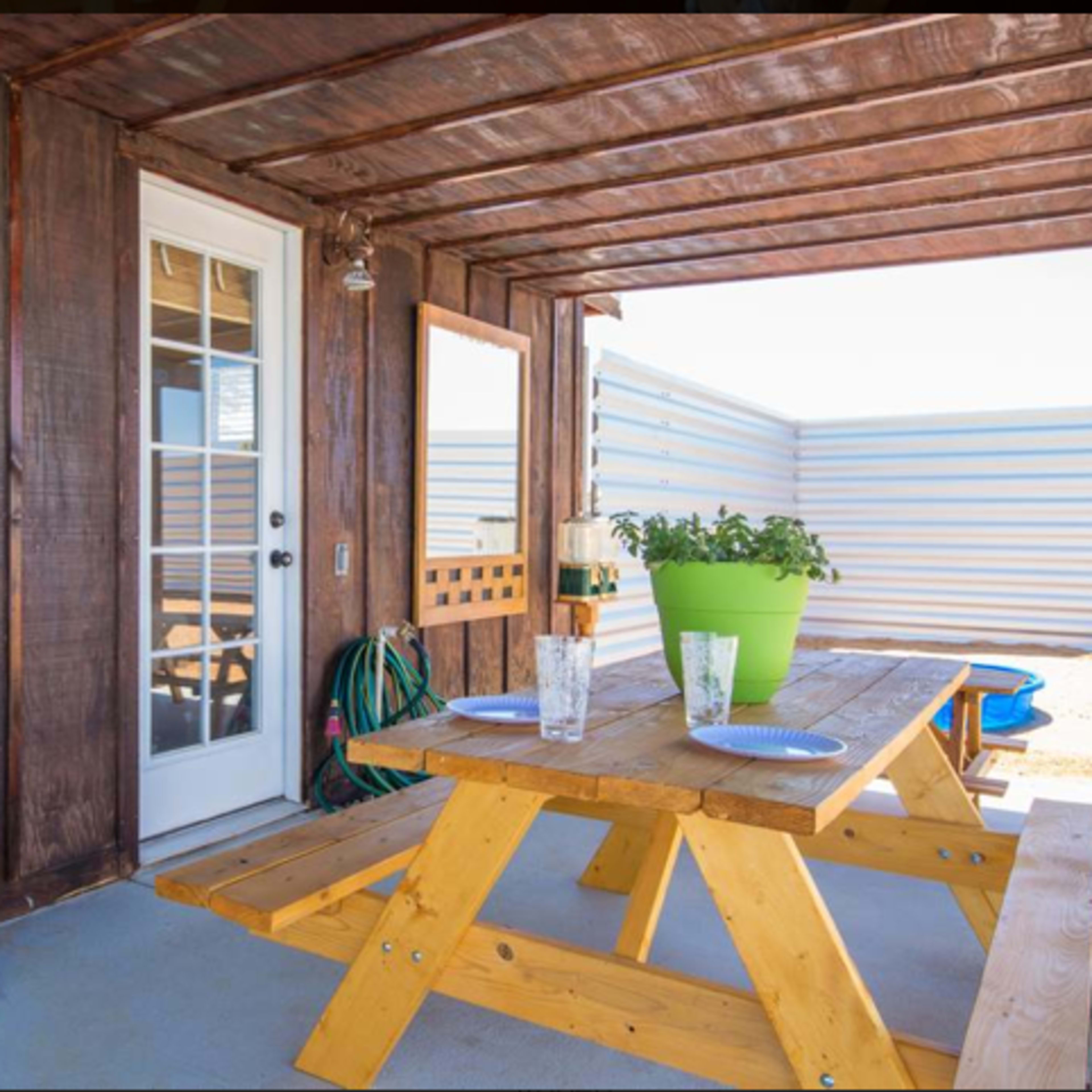 A wooden outdoor seating area features a picnic table with plates and glasses, beside a decorative wall with a sign and a plant.