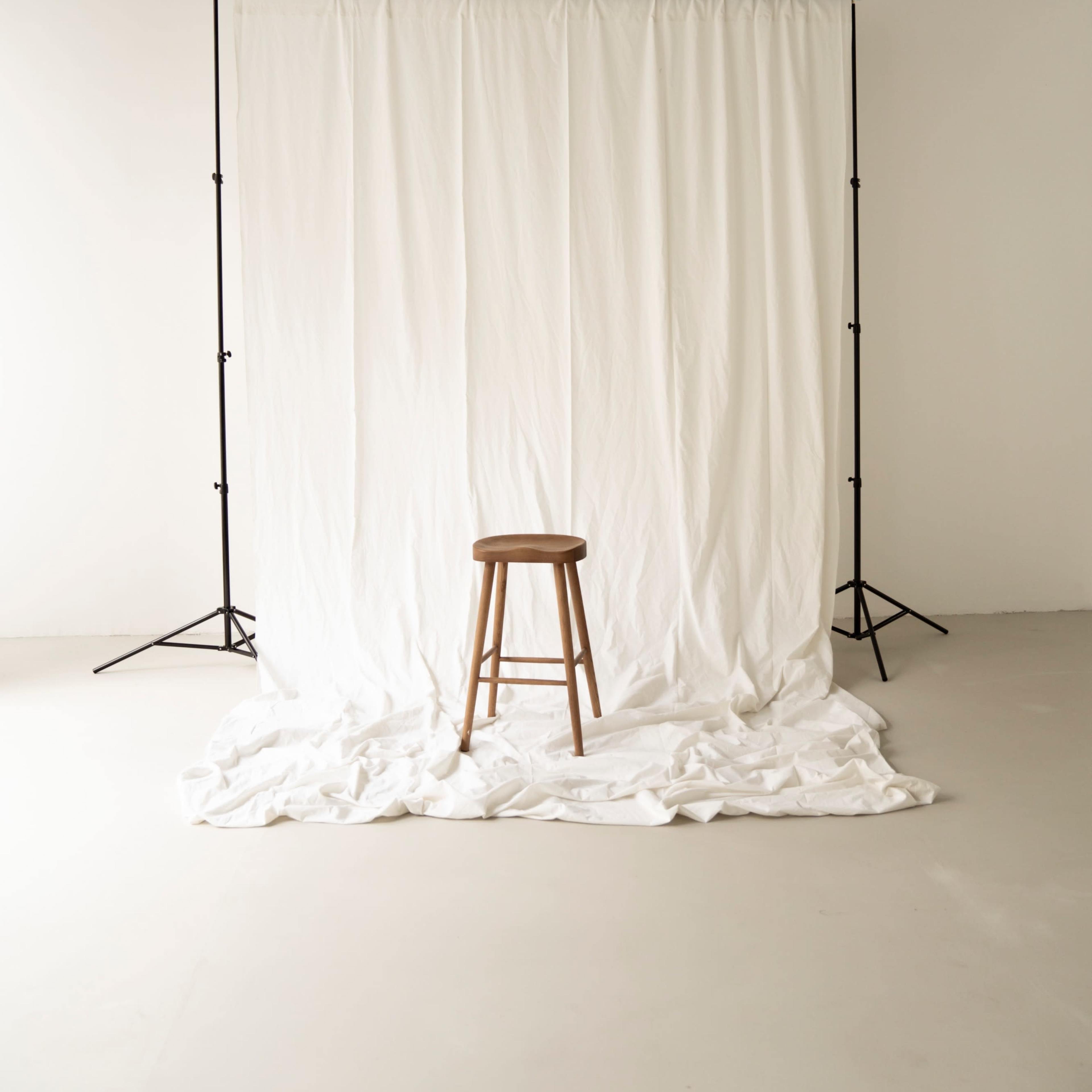 A wooden stool is positioned in front of a white backdrop draped on the floor.