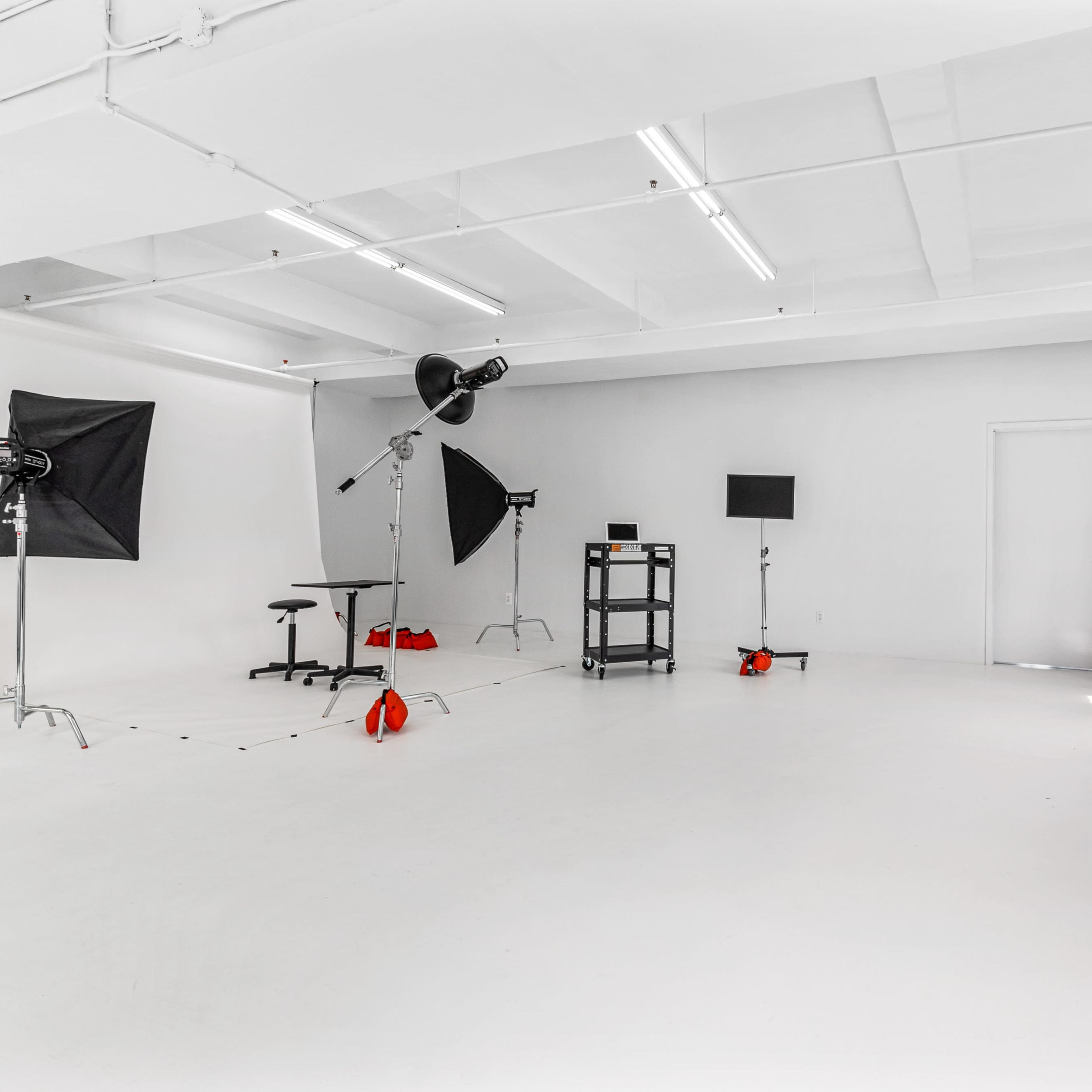 A well-lit photo studio features a white backdrop, various lighting equipment, a cart with equipment, and a dark couch against a wall.