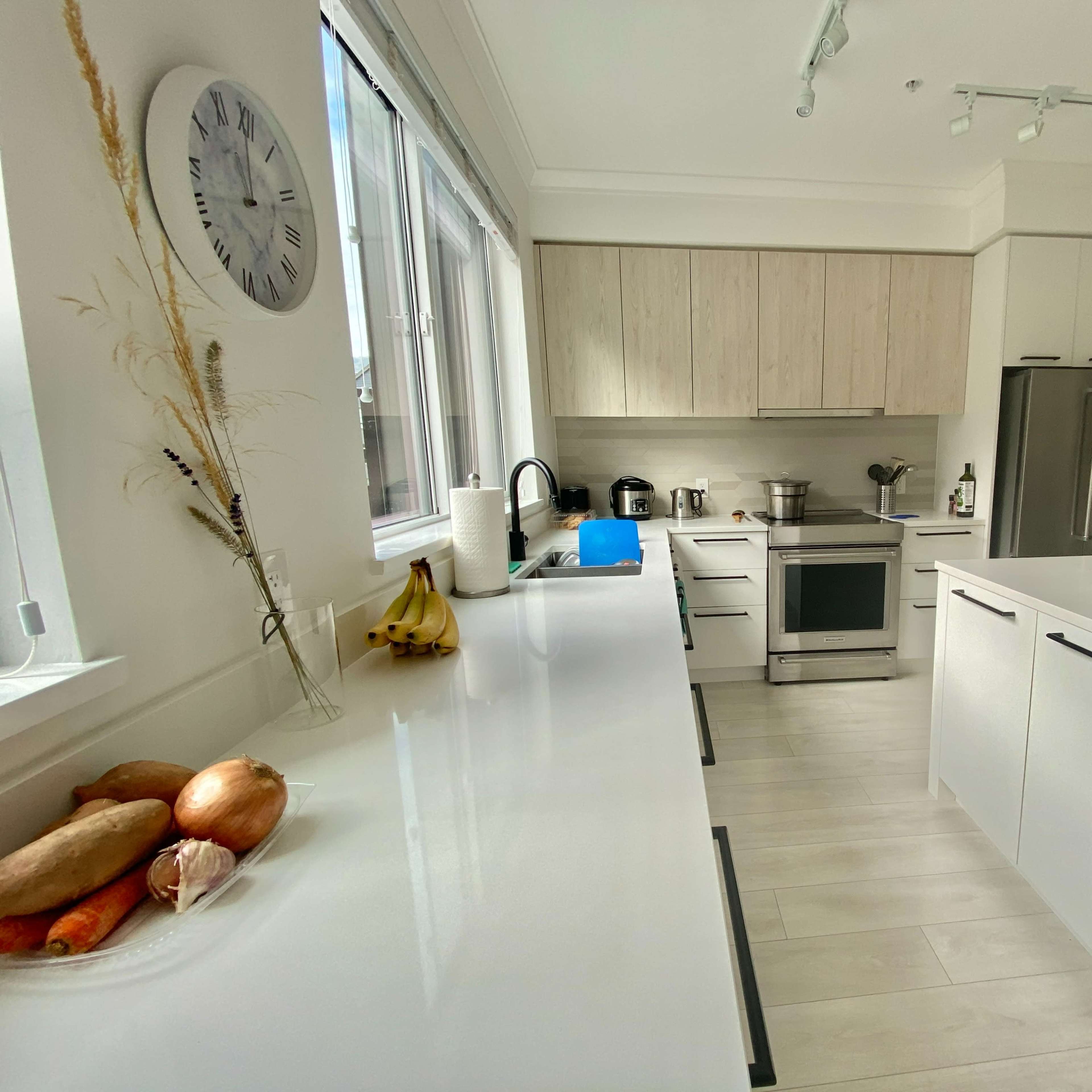 A modern kitchen features light wood cabinets, stainless steel appliances, and a white countertop with a bowl of fruits.