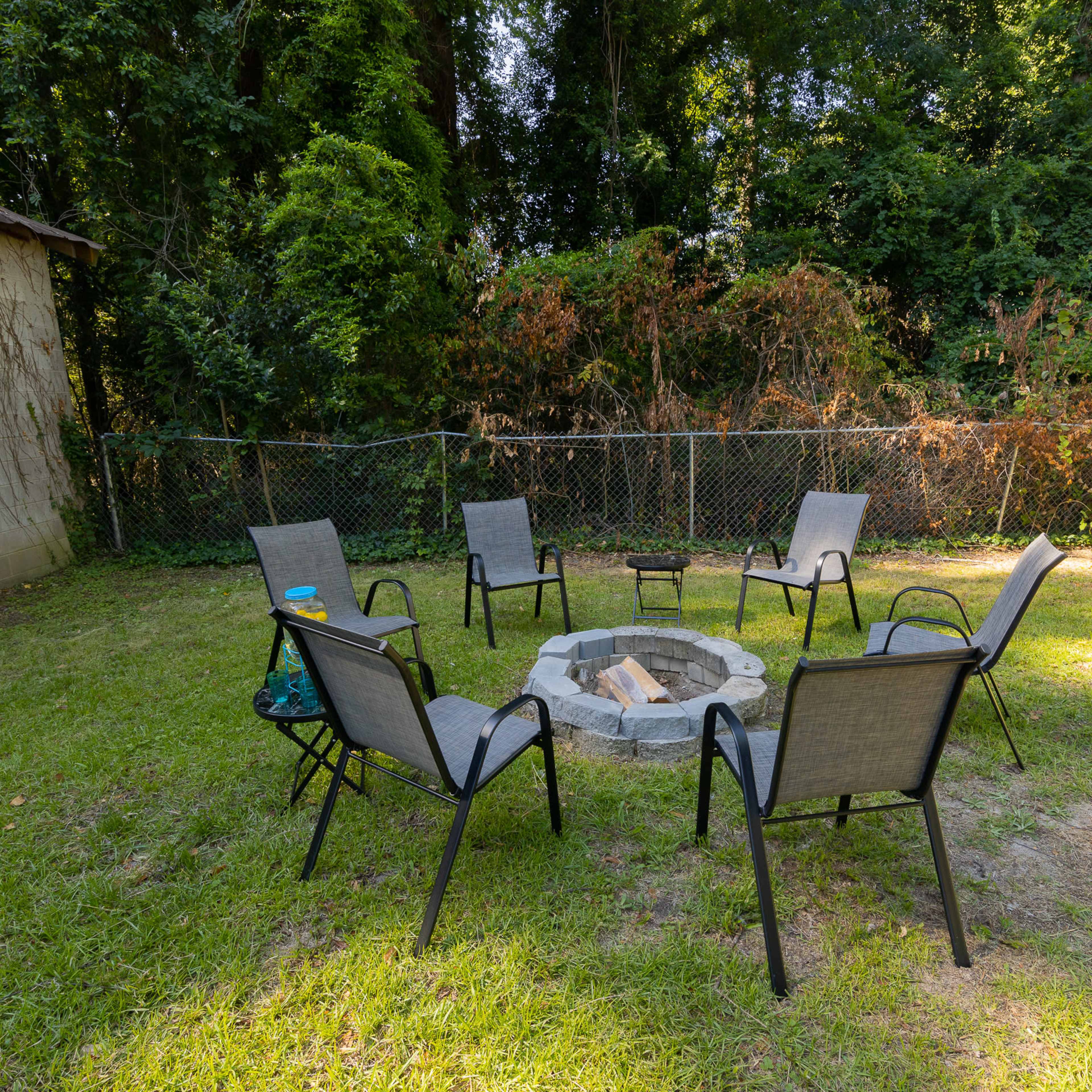 A circular stone fire pit is surrounded by six chairs in a grassy backyard, with a fence and trees in the background.