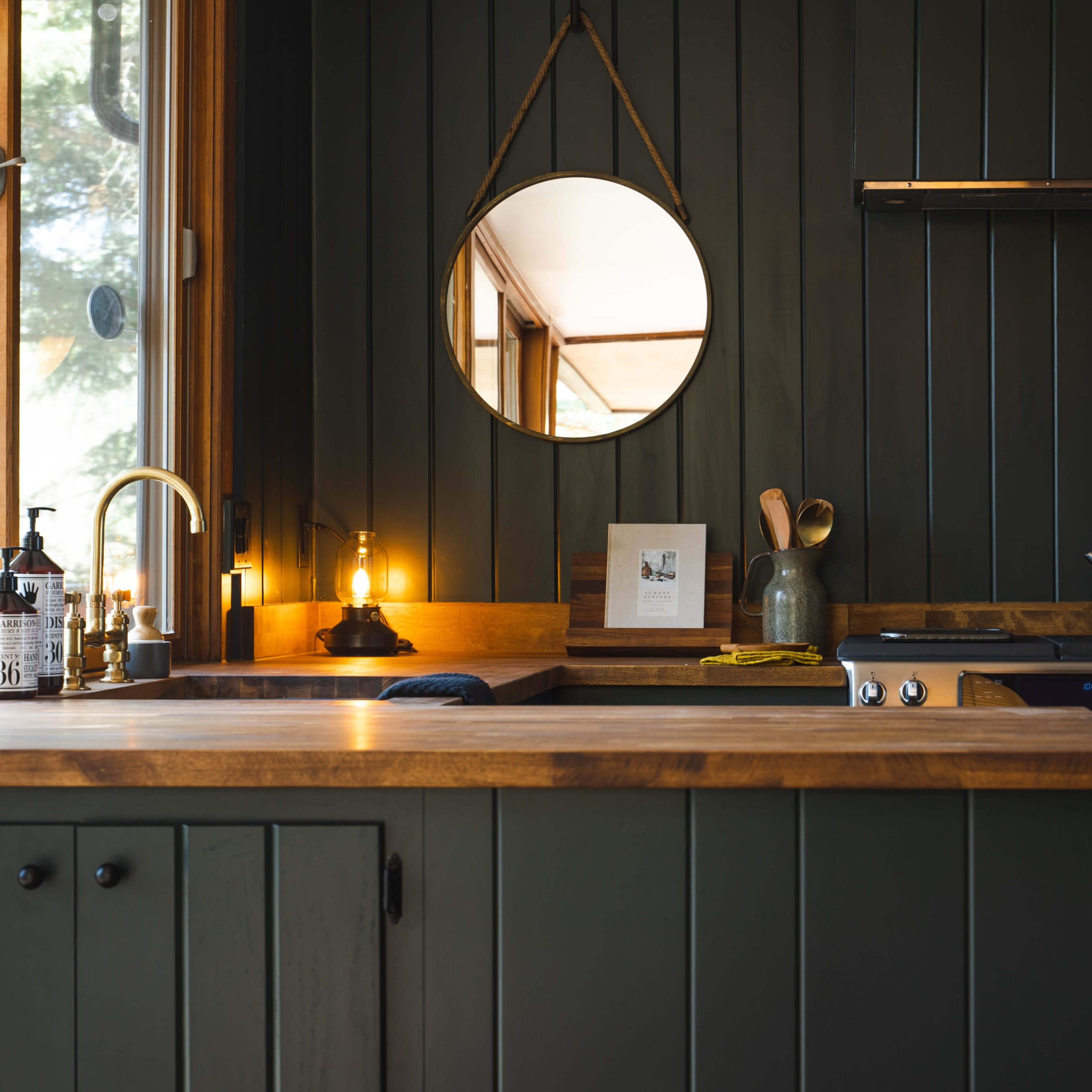 A modern kitchen with dark green paneling, a wooden countertop, and a round mirror mounted on the wall.