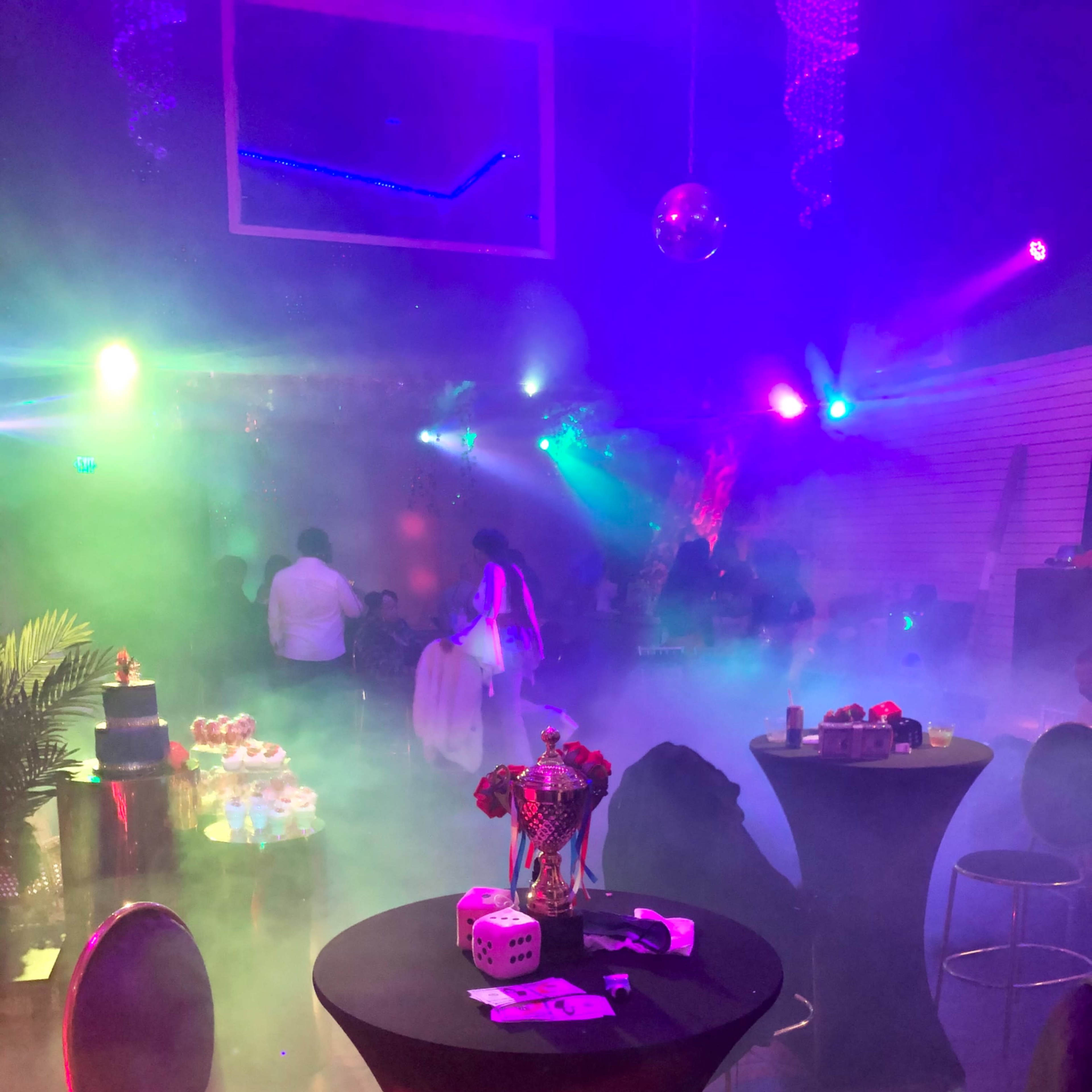 A dimly lit party scene features tables with decorated snacks and a foggy atmosphere enhanced by colorful lighting and silhouettes of people mingling.