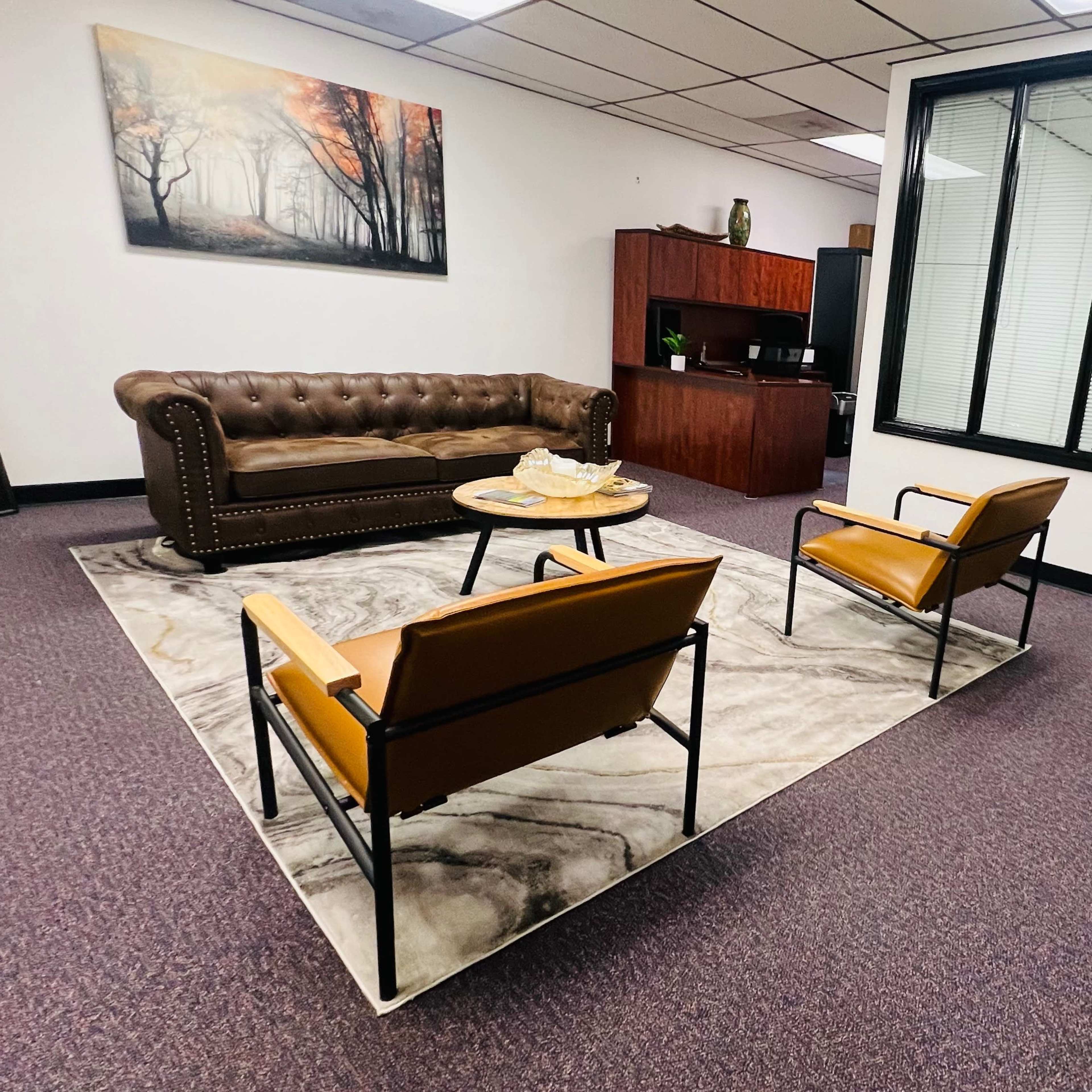 A professional office waiting area with a brown leather sofa, two orange chairs, a coffee table on a patterned rug, and a glass-walled office in the background.