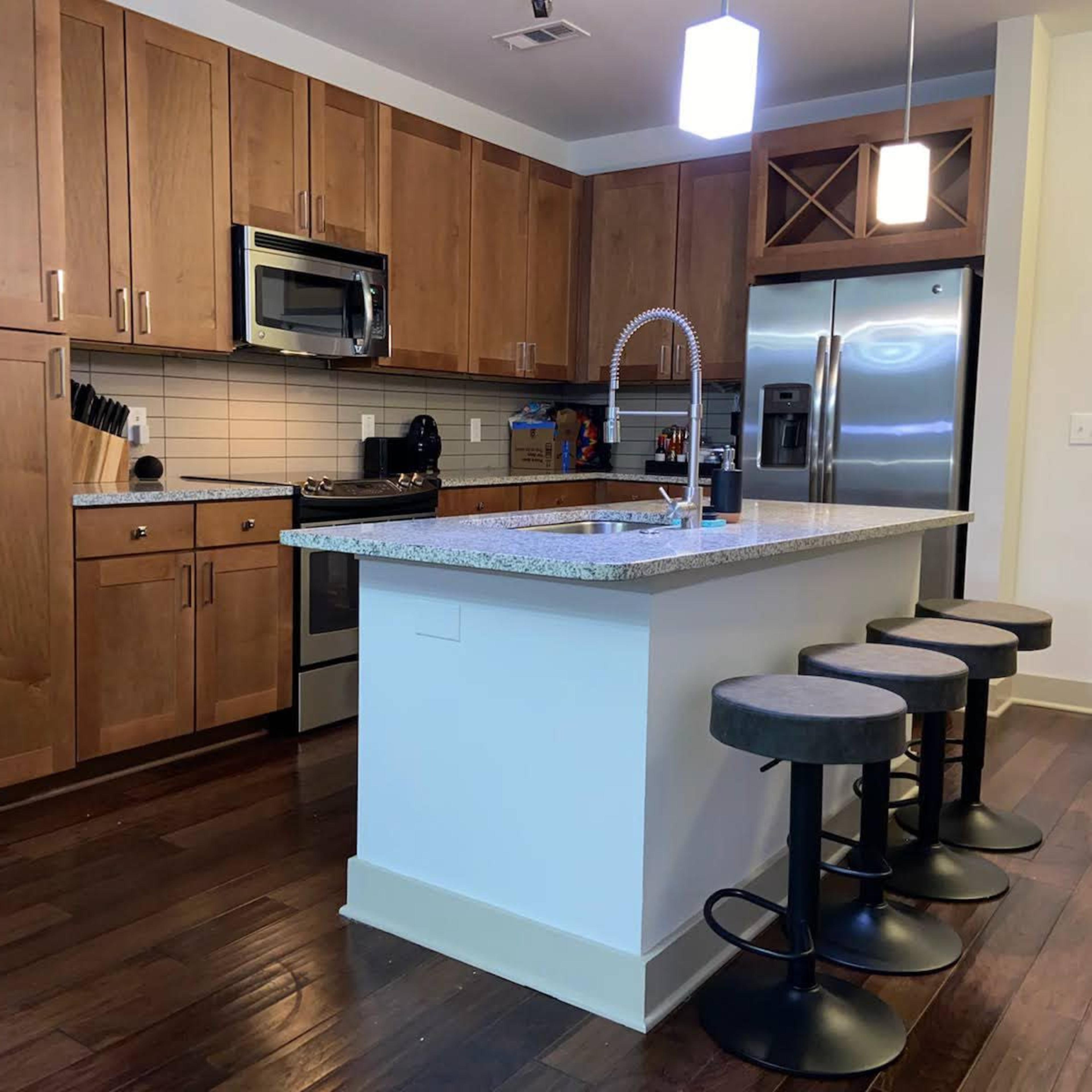 A modern kitchen features wooden cabinets, stainless steel appliances, a granite countertop island with three stools, and warm wooden flooring.
