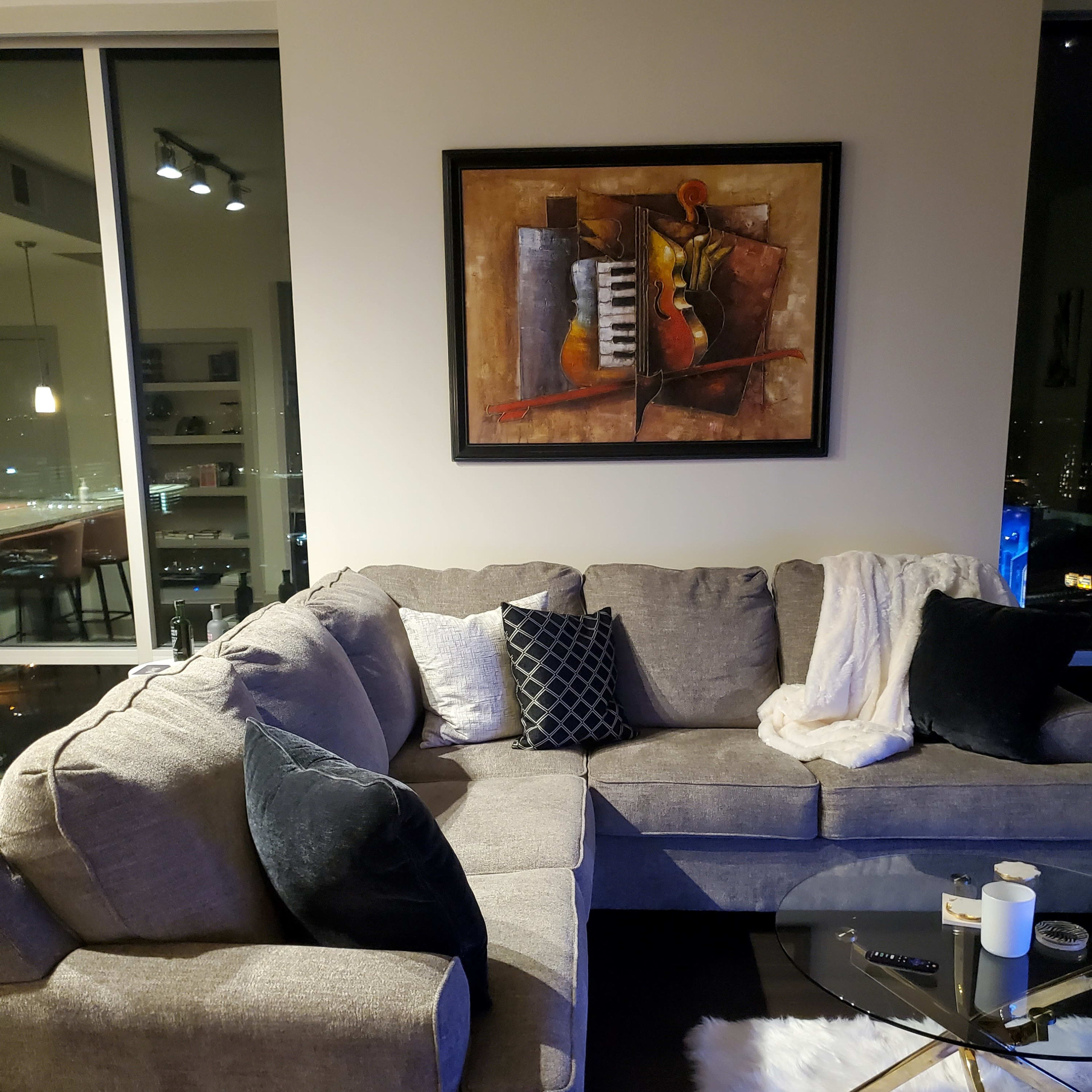 A gray sectional sofa with black and white pillows, accompanied by a round coffee table on a white rug, and a colorful abstract painting hanging on the wall.