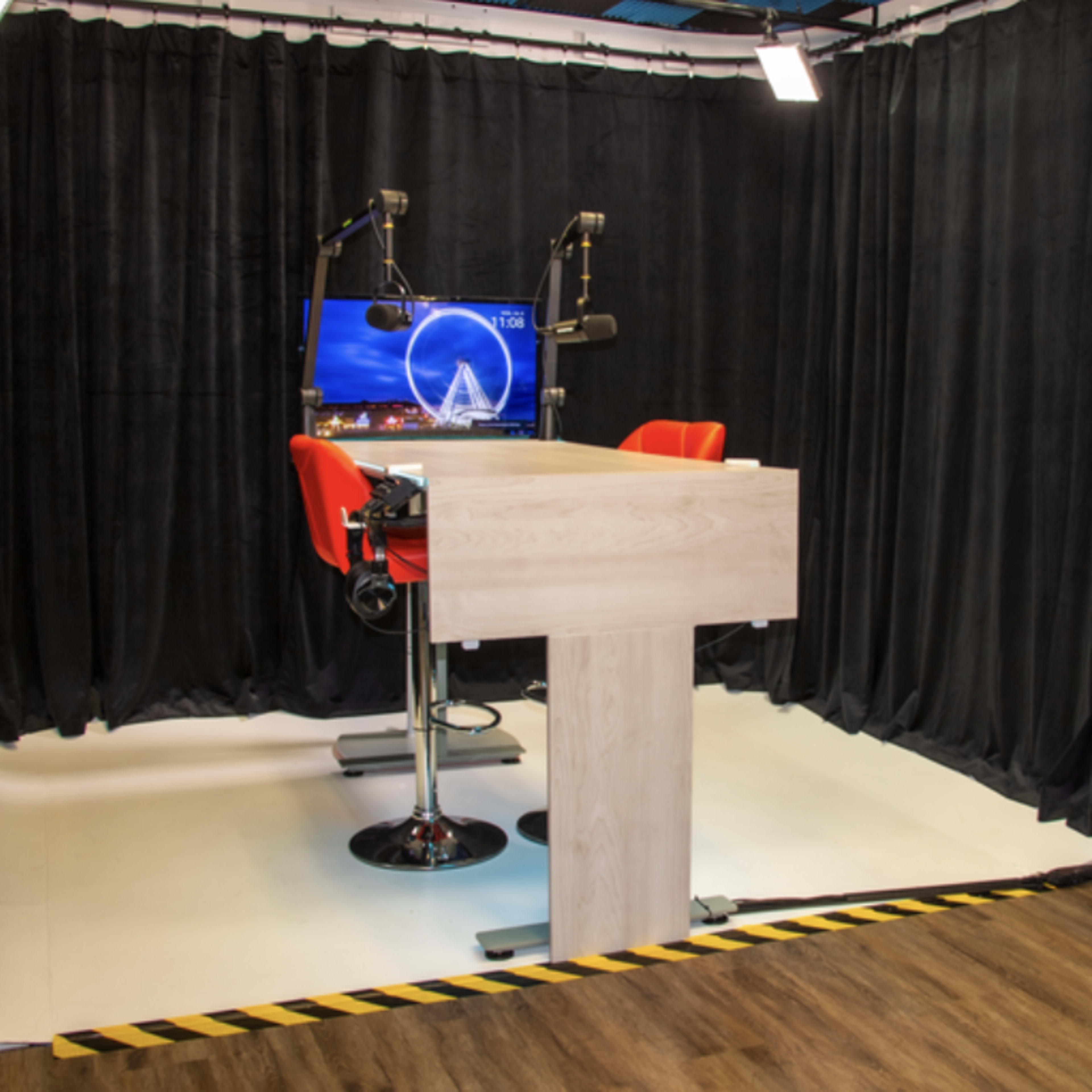 A recording studio with a central table, two red swivel chairs, and a monitor, surrounded by black curtains and a patterned blue wall.