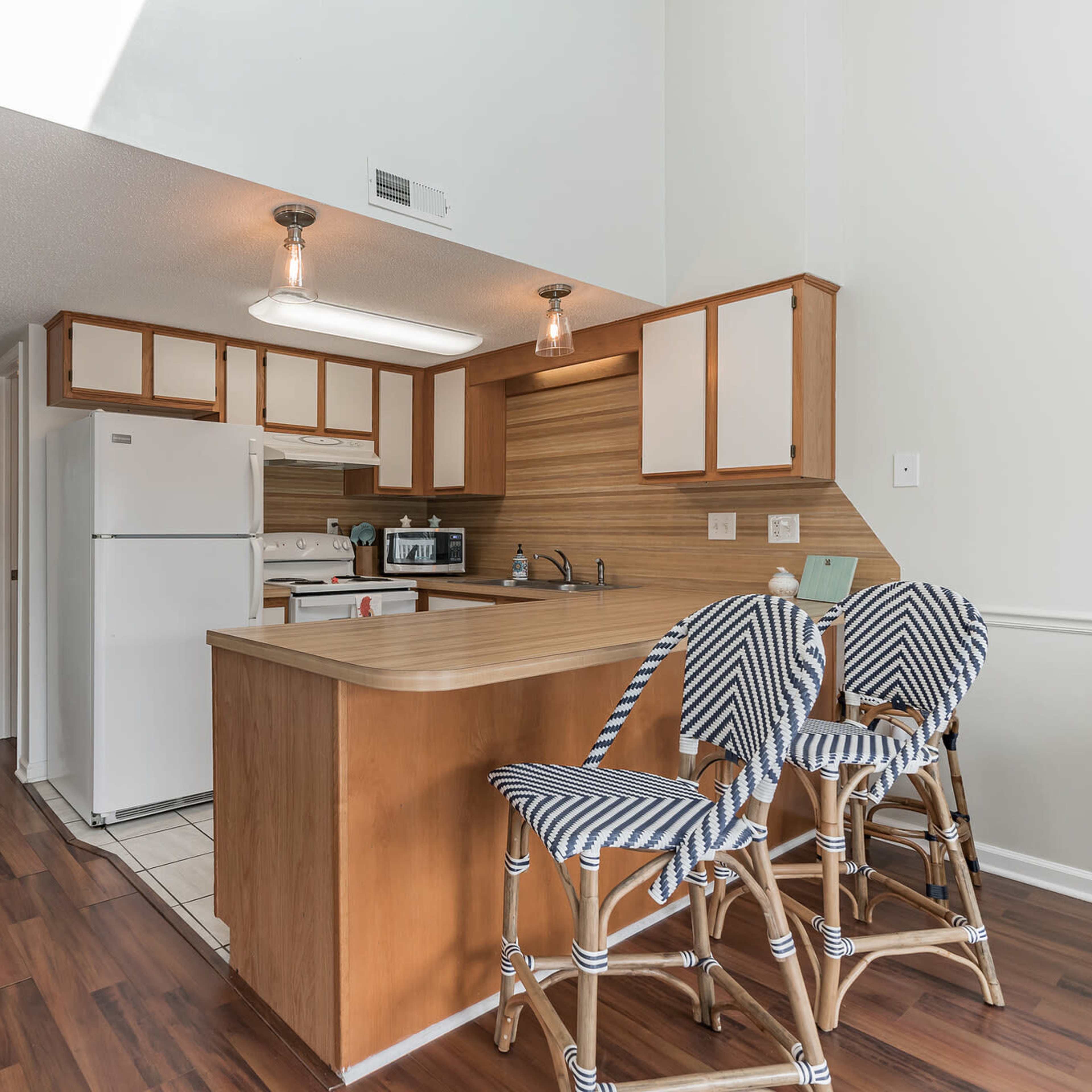 A modern kitchen with a light-colored wooden bar, striped stools, and white appliances, leading to a hallway in the background.