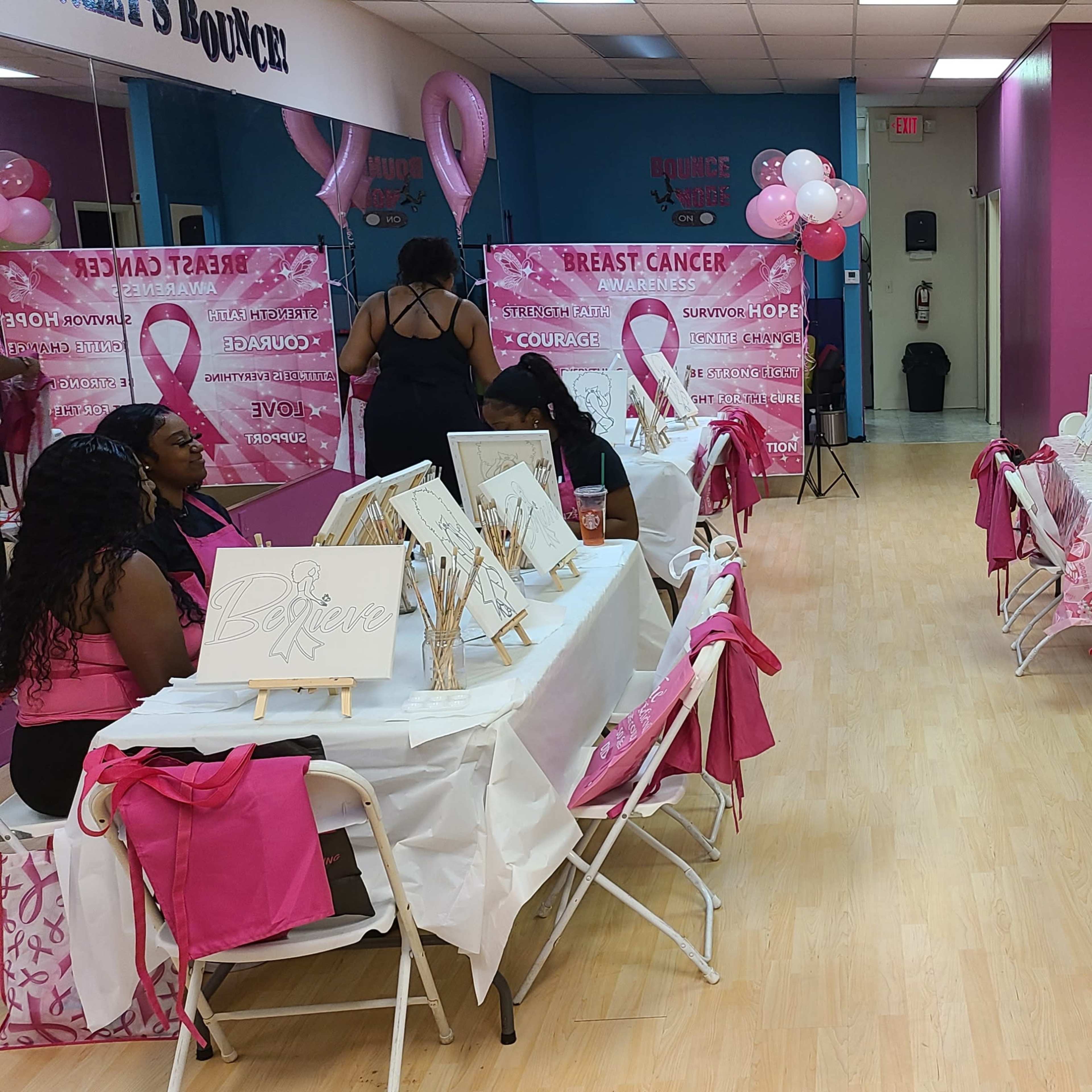 A brightly colored room set up for a breast cancer awareness event, featuring tables with easels and blank canvases, surrounded by decorations in pink.