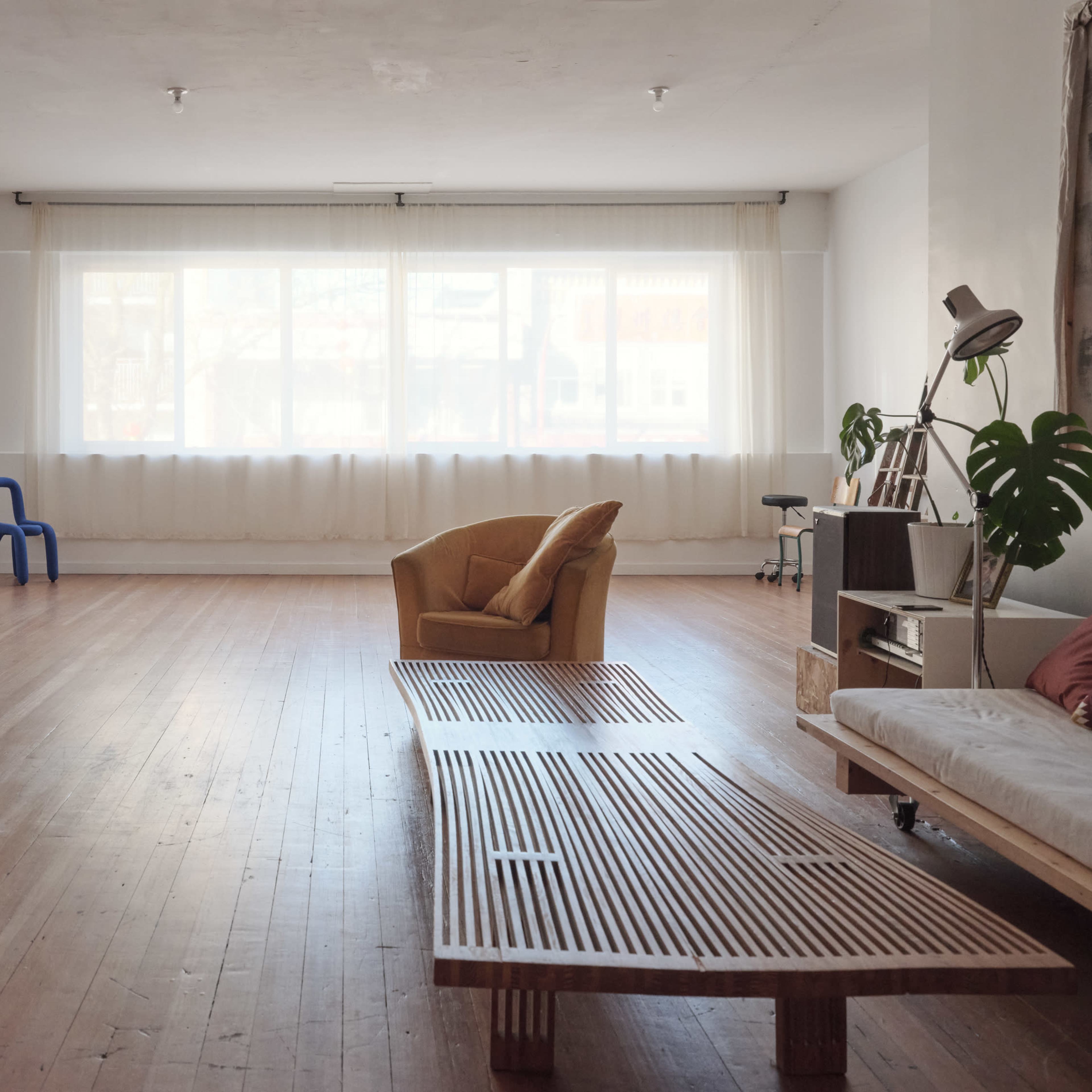 A spacious room with wooden floors, large windows, and minimalist furniture, including a yellow armchair, a wooden coffee table, and a blue chair.
