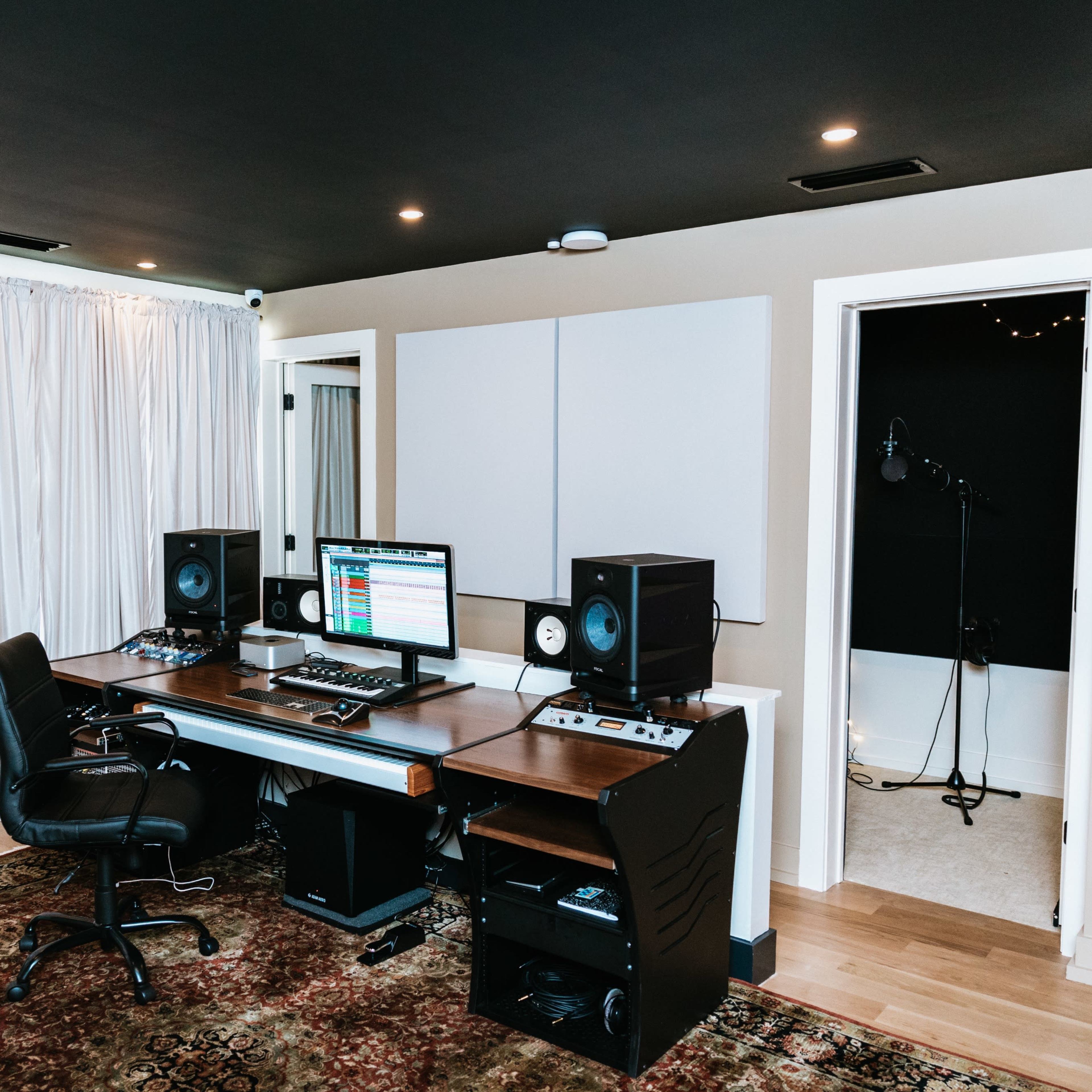 A music production studio featuring a desk with computer monitors, studio speakers, and a keyboard, along with soundproof panels and a microphone setup in the recording area.