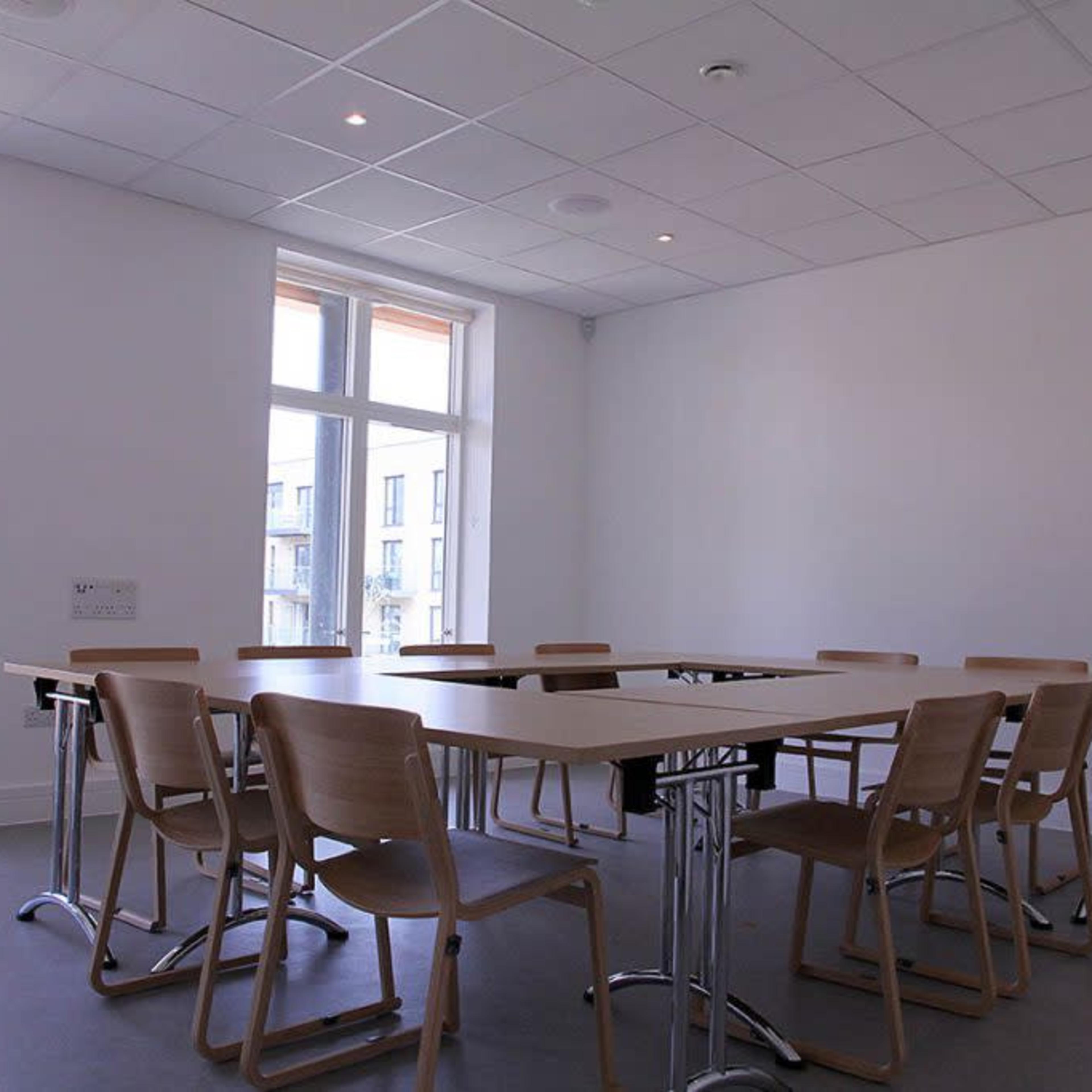 A minimalist conference room features a rectangular table surrounded by eight wooden chairs, with large windows letting in natural light.