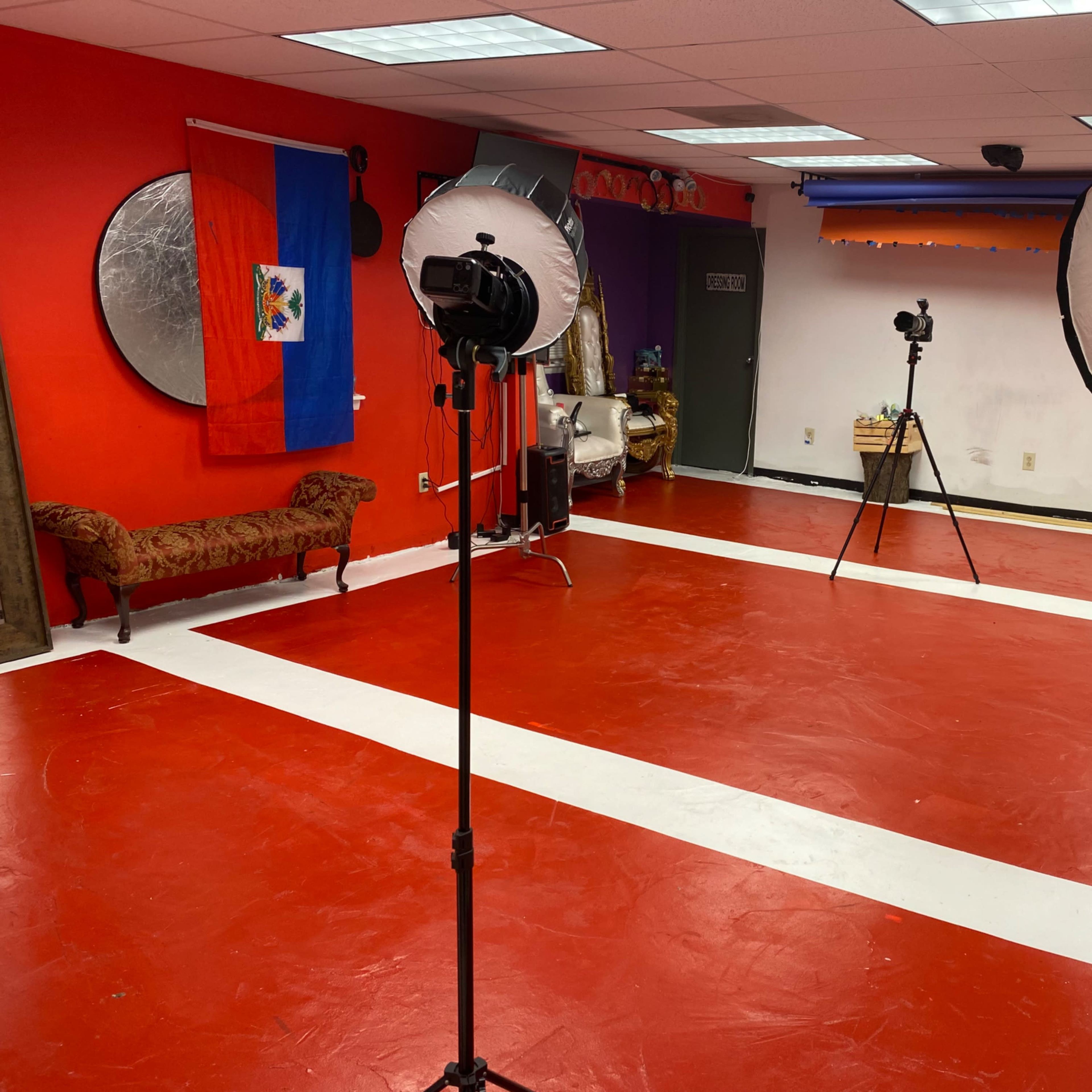 An empty studio space with red flooring, white stripes, two large softbox lights on stands, and a bench, along with a Haitian flag hanging on the wall.