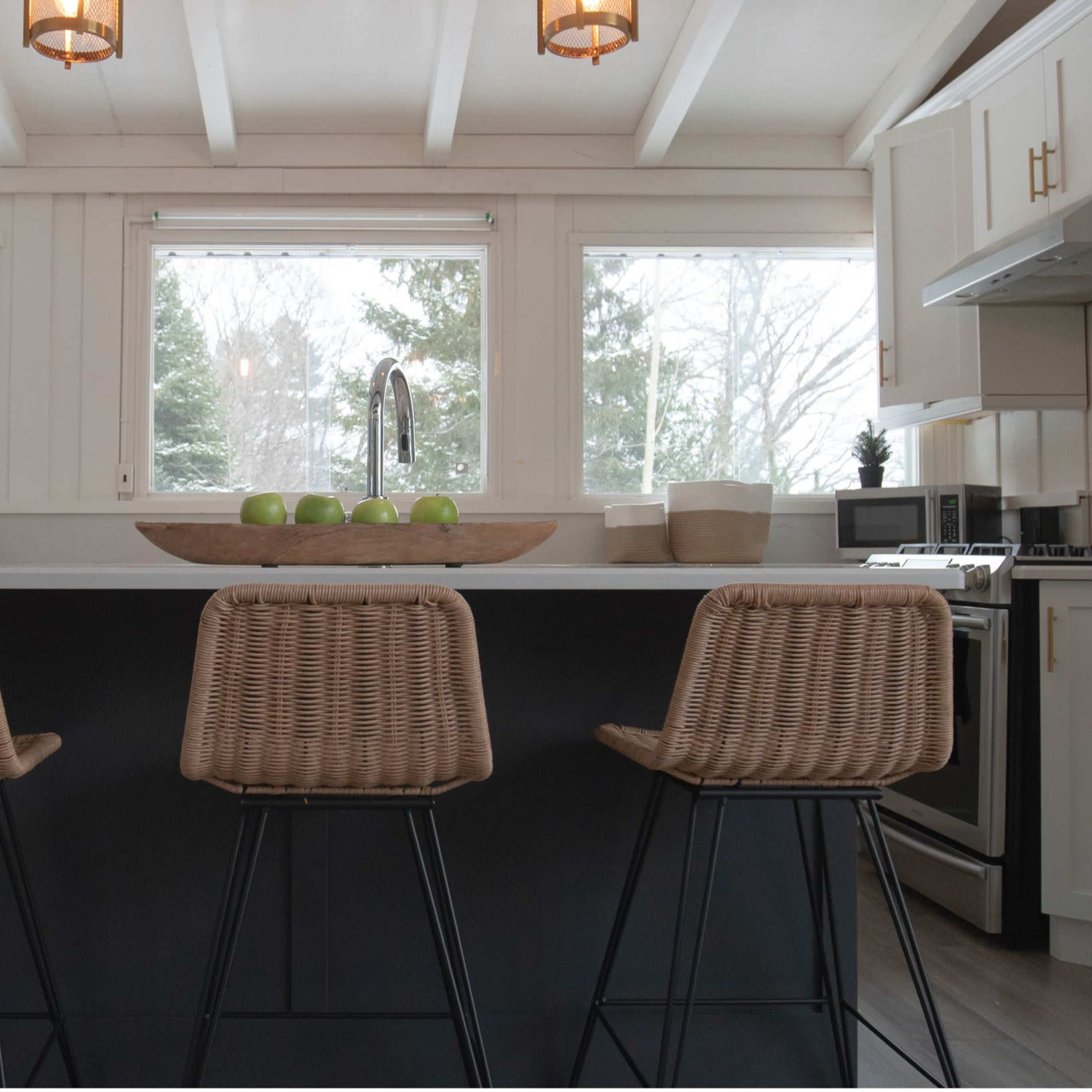 A modern kitchen features an island with three woven bar stools, surrounded by large windows and equipped with stainless steel appliances.