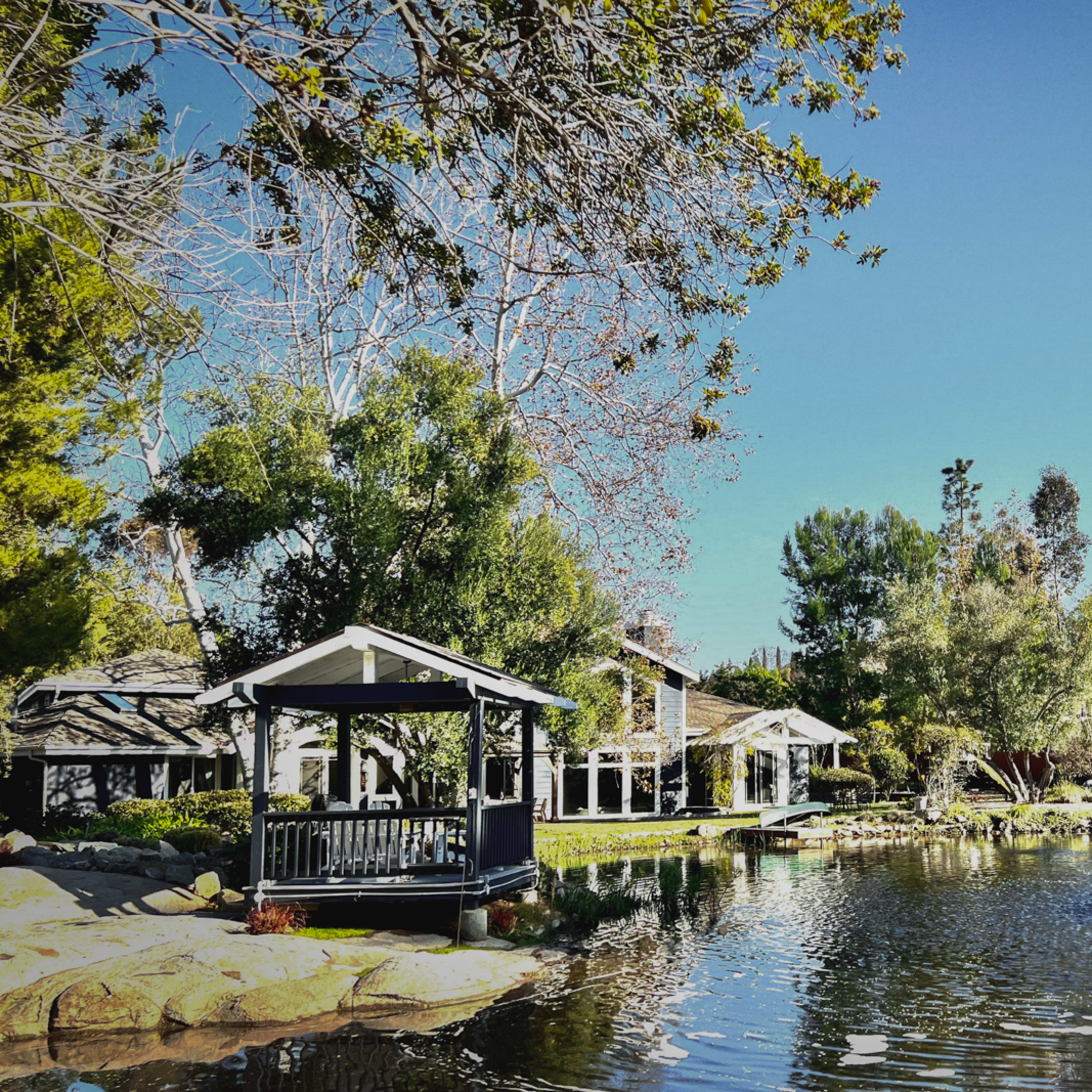 A gazebo and building are situated next to a pond featuring a fountain, surrounded by trees and shrubs.