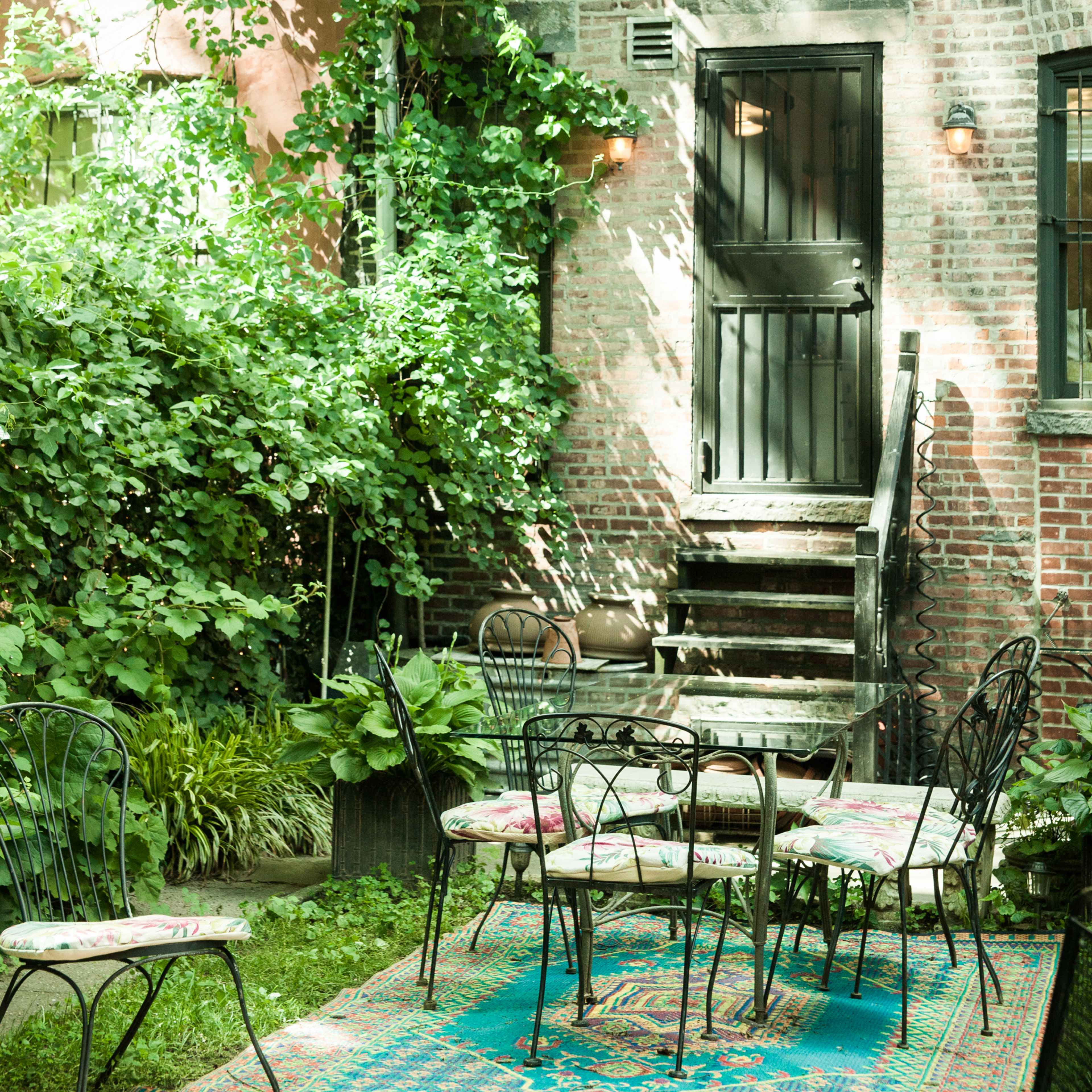 A garden area features a set of six wrought-iron chairs arranged around a table on a colorful rug, with a brick wall and a wooden door in the background.