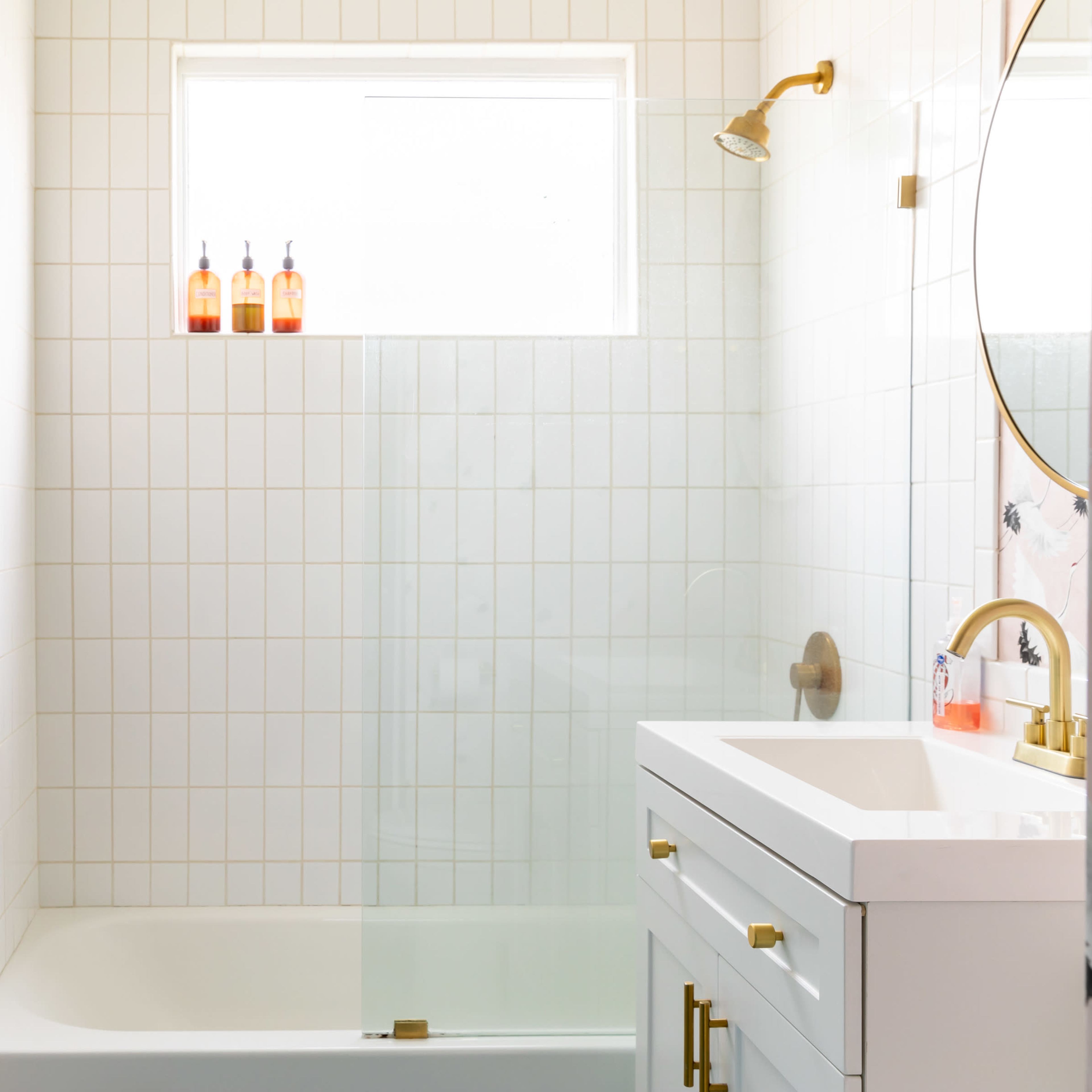 A modern bathroom featuring a bathtub with a glass panel, a white vanity with gold fixtures, and a window above the tub displaying three bottles.