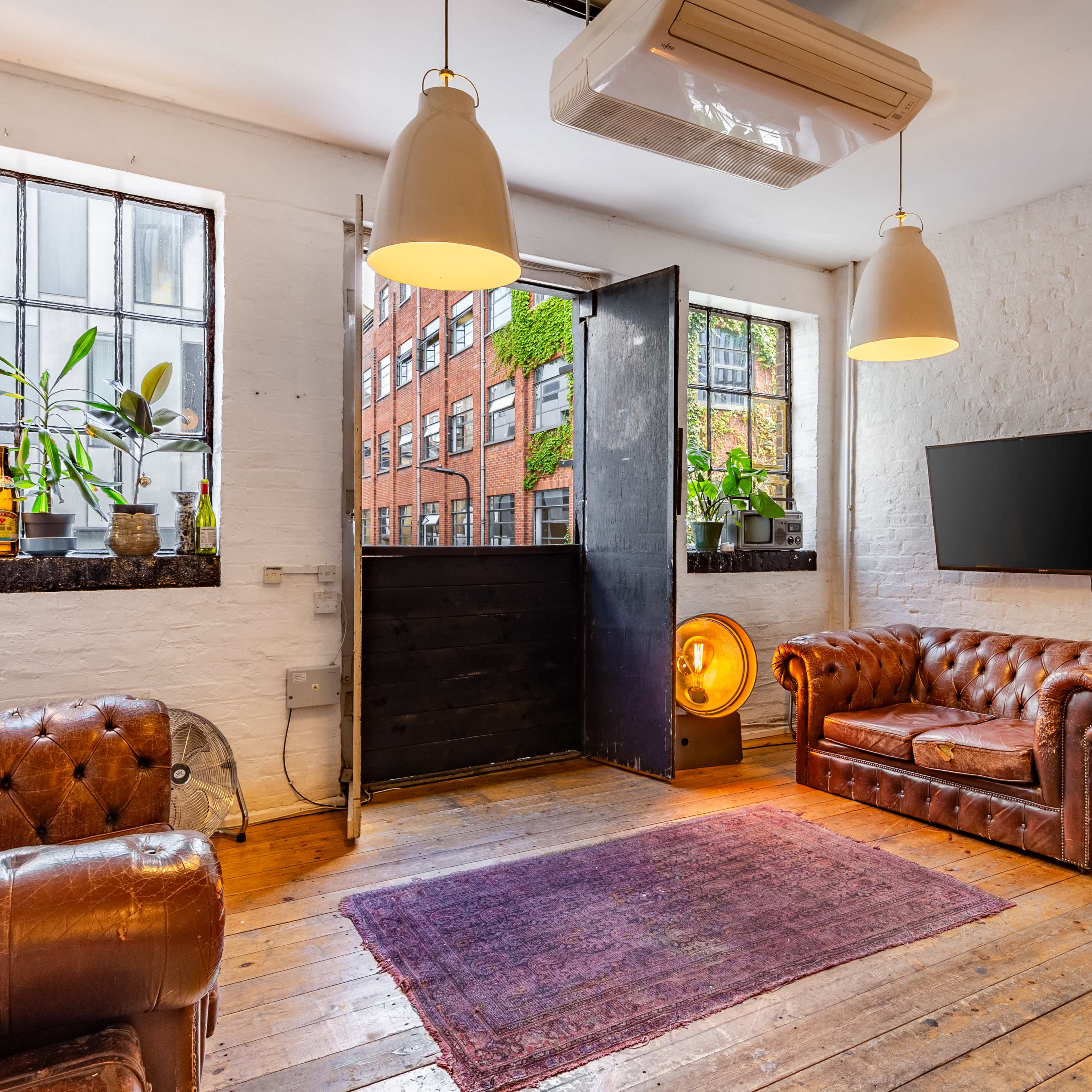 The room features two brown leather sofas, a TV mounted on the wall, and large windows allowing light to stream in, with a door leading outside.