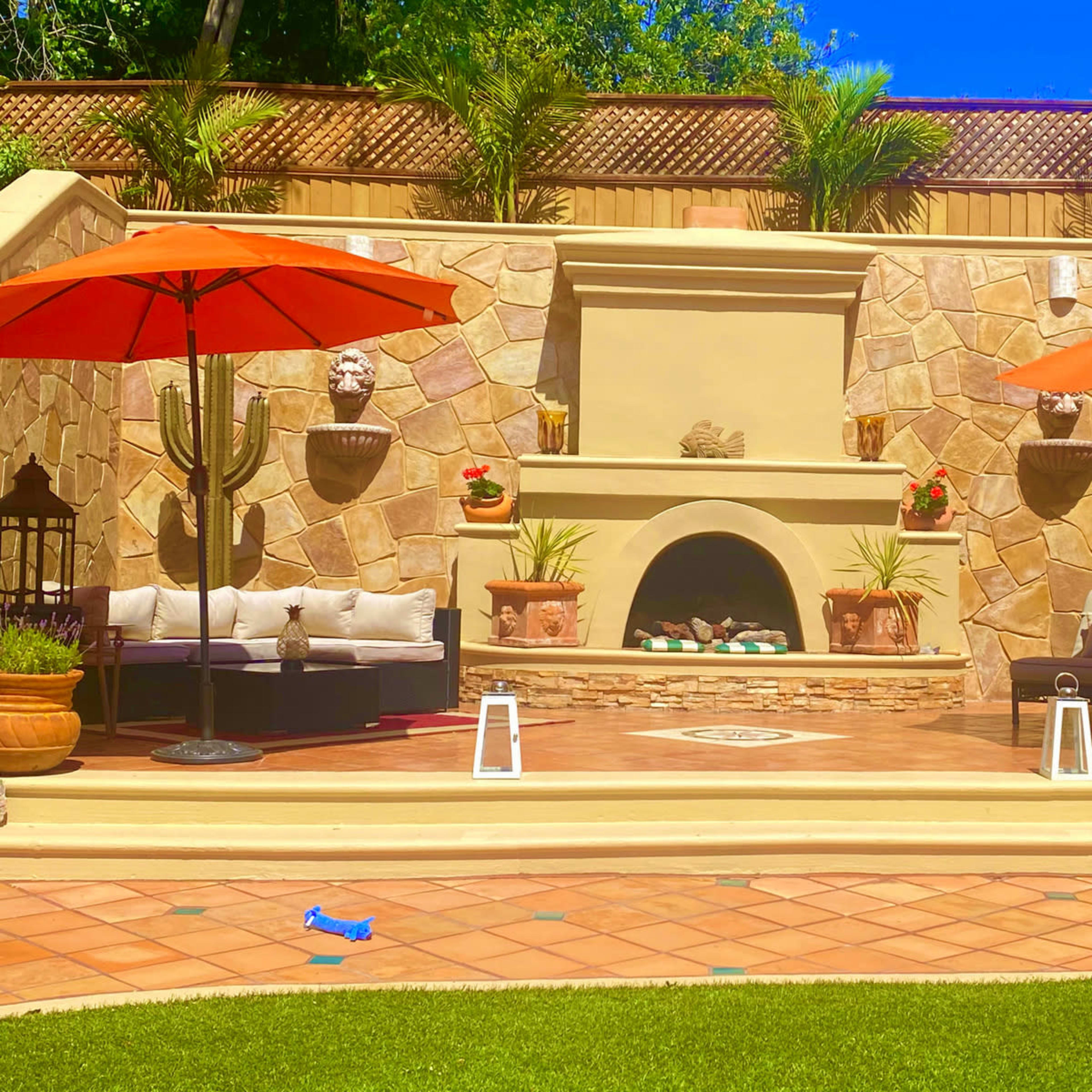 The outdoor patio features a stone wall with a fireplace and two red umbrellas shading seating areas, surrounded by potted plants and decorative accents.