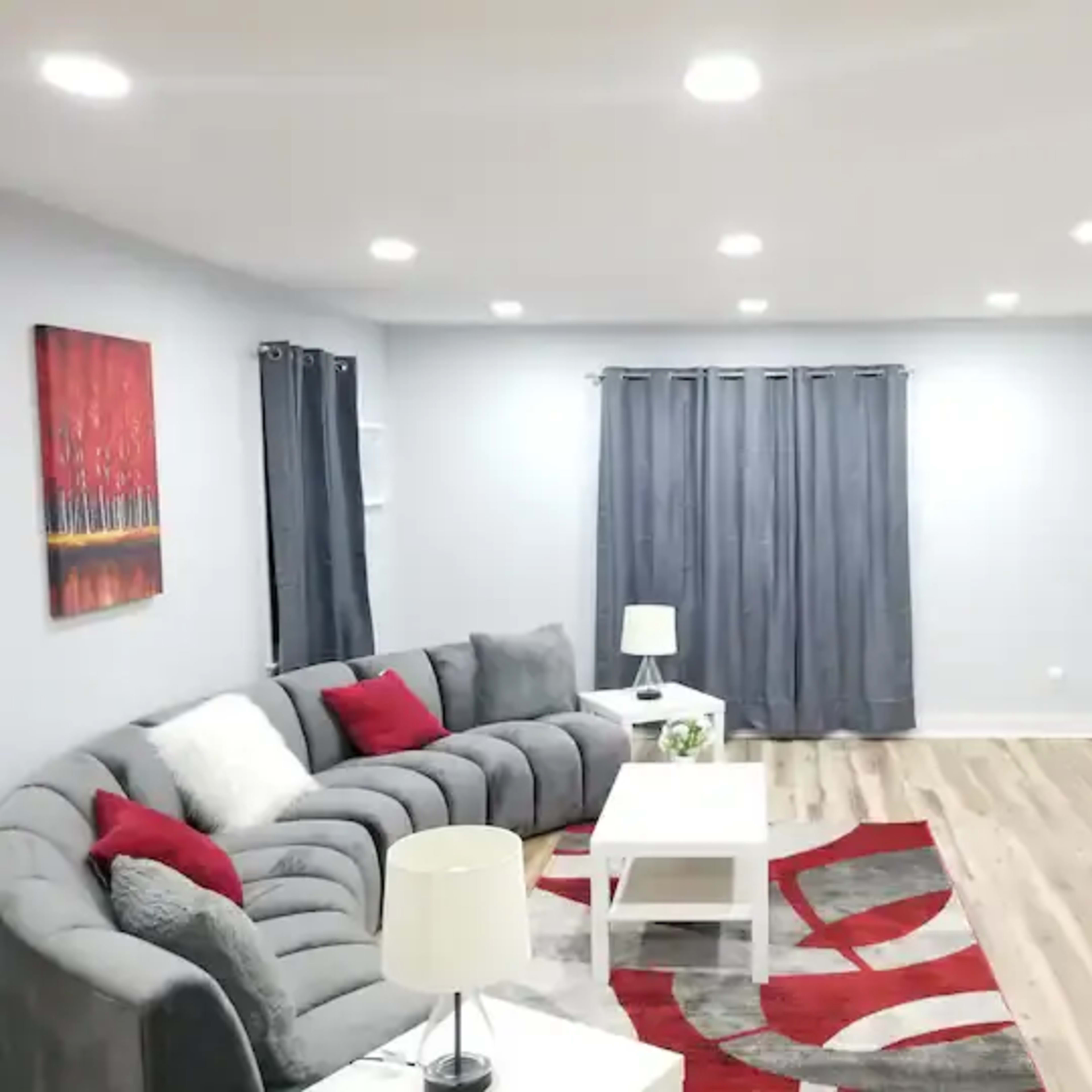 A modern living room features a gray sectional sofa, a white coffee table, a wall-mounted television, and decorative curtains, with a red and gray area rug on the wooden floor.
