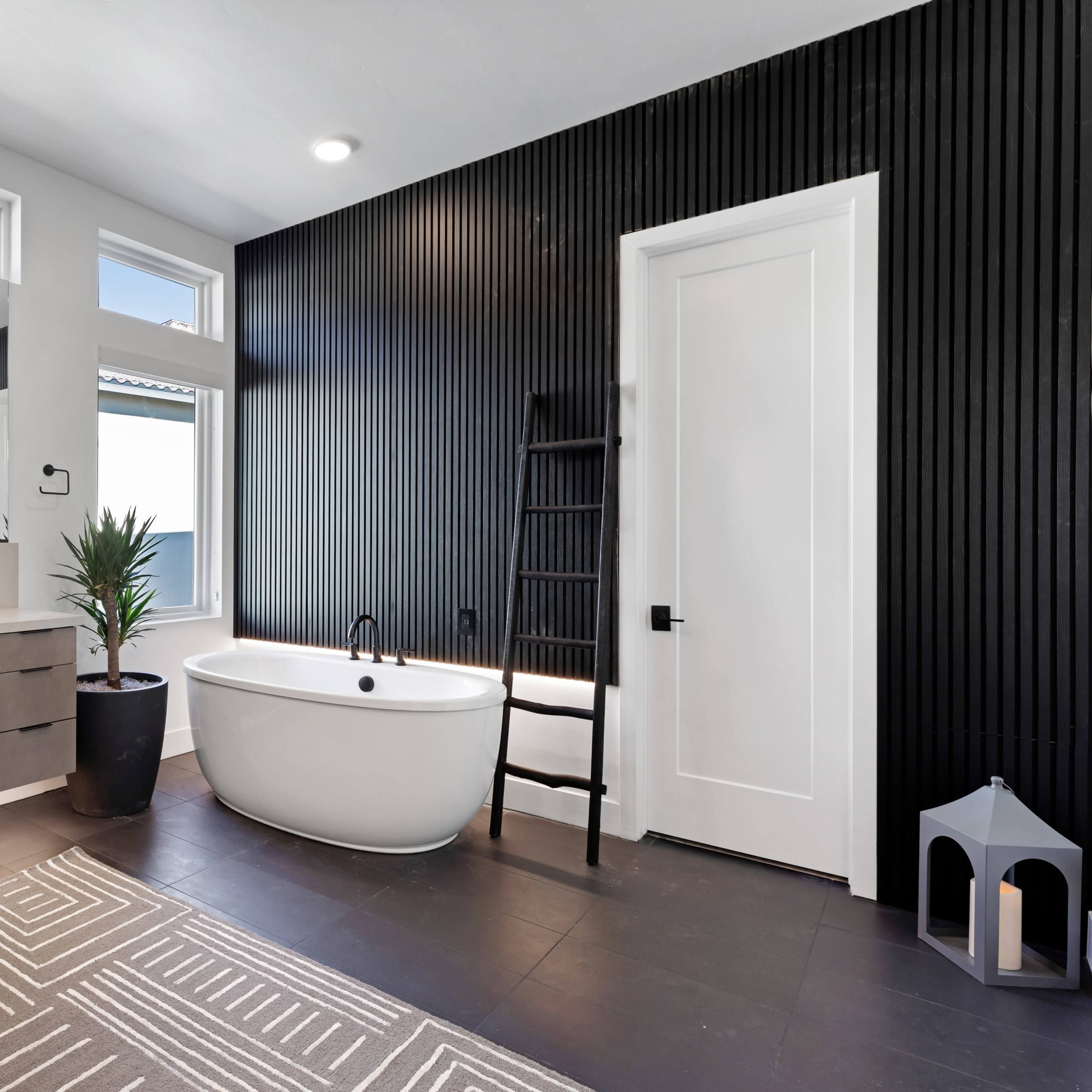 A modern bathroom featuring a freestanding bathtub, a ladder leaning against a textured black wall, and a vanity with drawers.