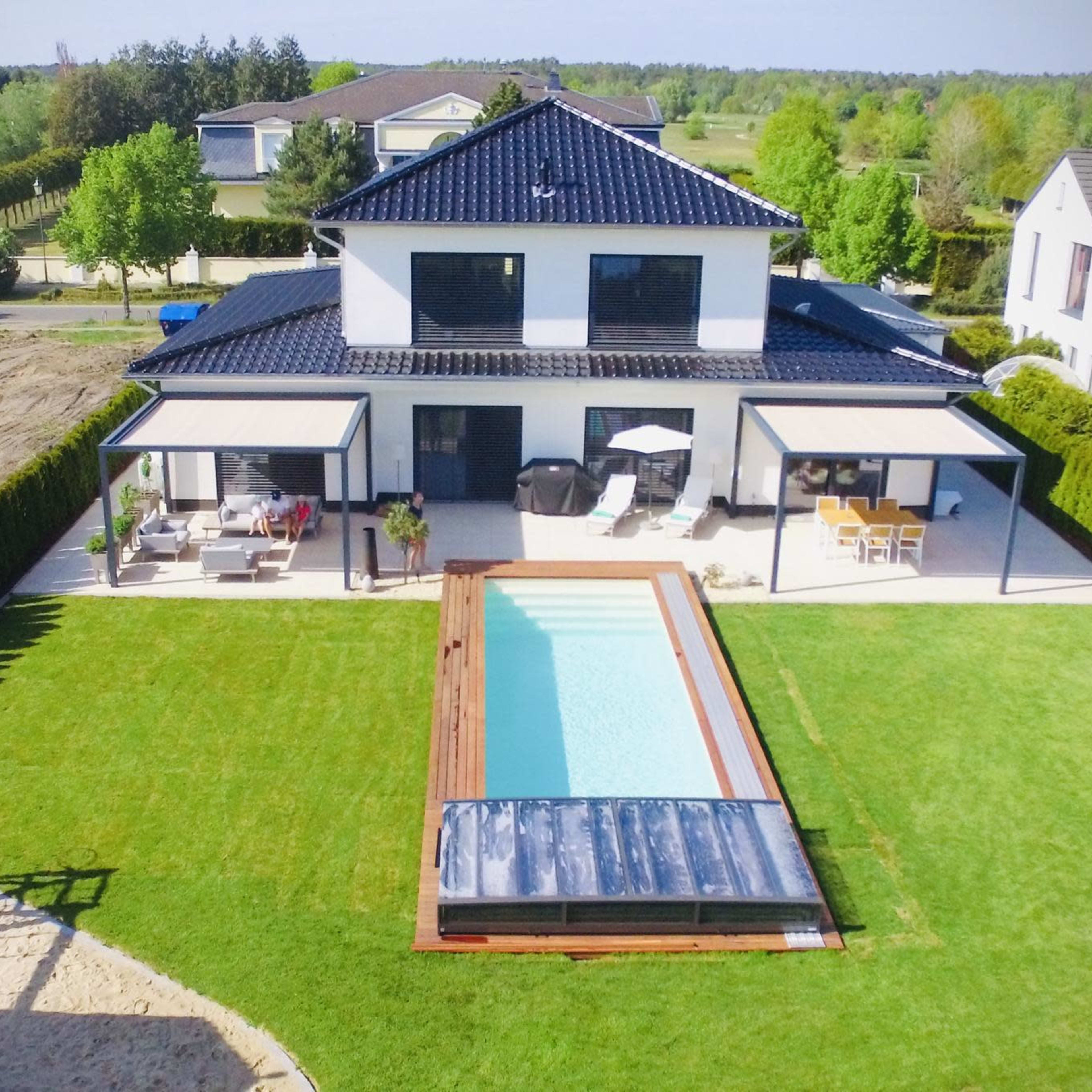 A modern two-story house with a pool and outdoor seating area is surrounded by a grassy lawn and trees.