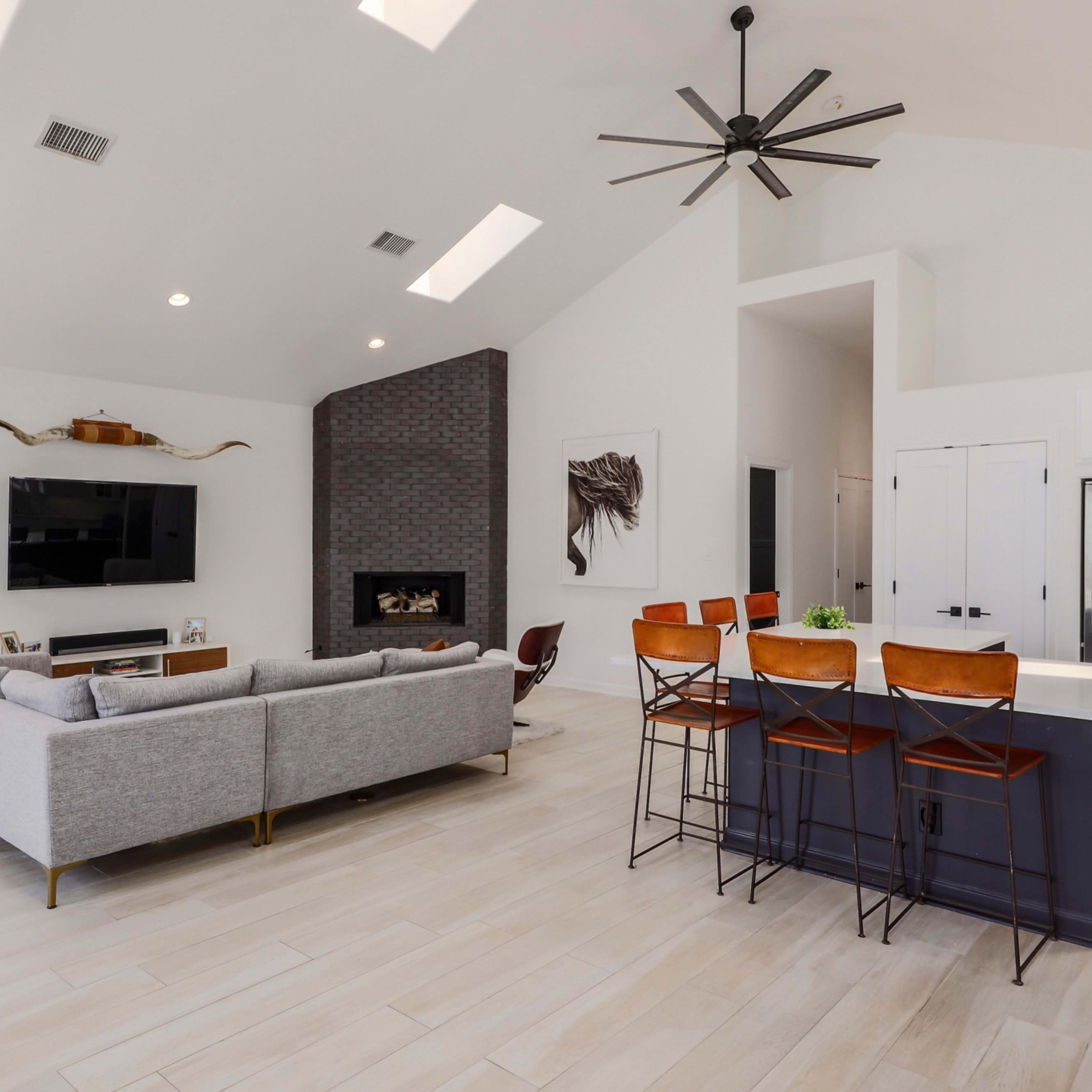 A modern living room and kitchen area with a gray sectional sofa, a flat-screen TV mounted on the wall, a dark brick fireplace, and a kitchen island with bar stools.