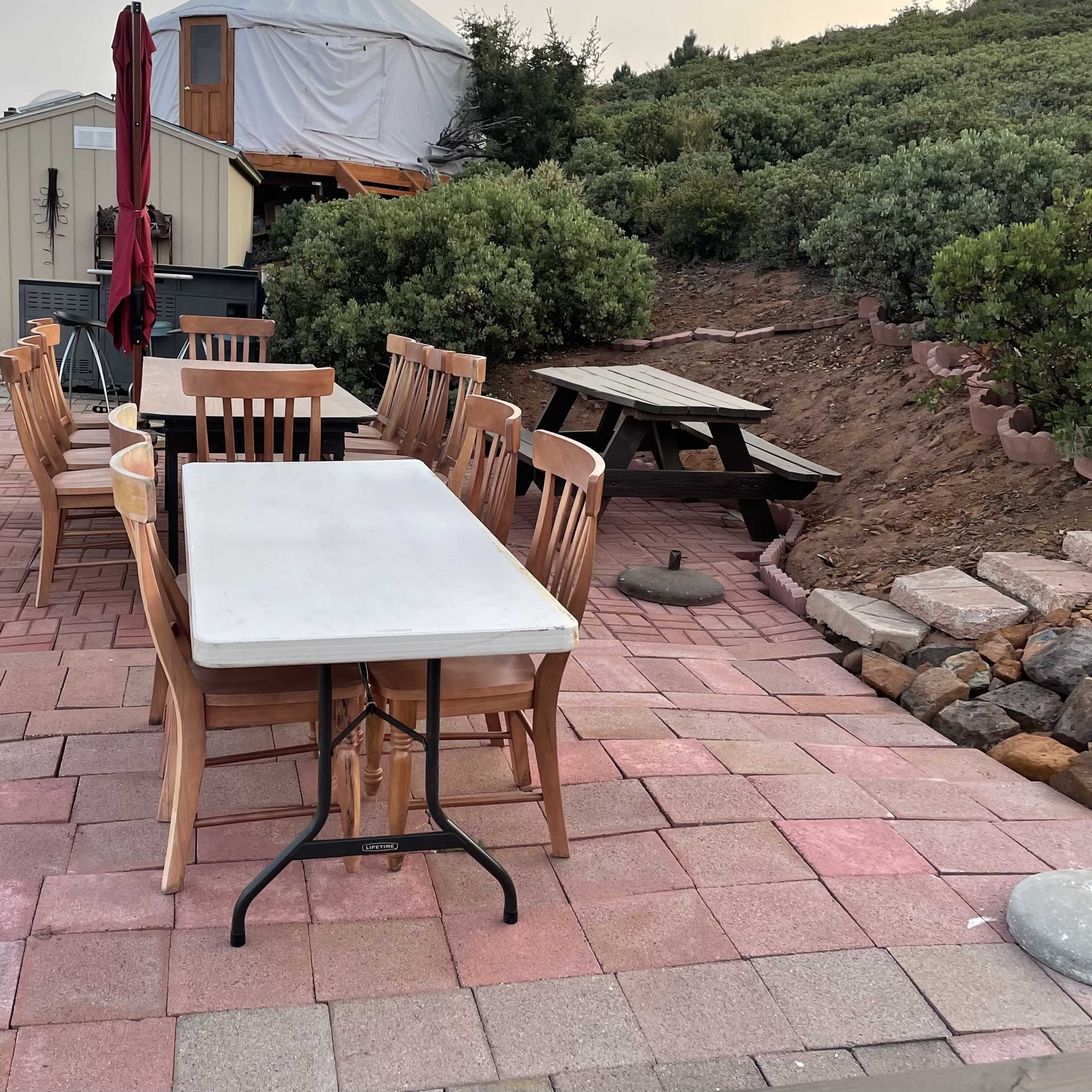 A rectangular white table is surrounded by wooden chairs on a paved patio, with a picnic table and a shaded area in the background.