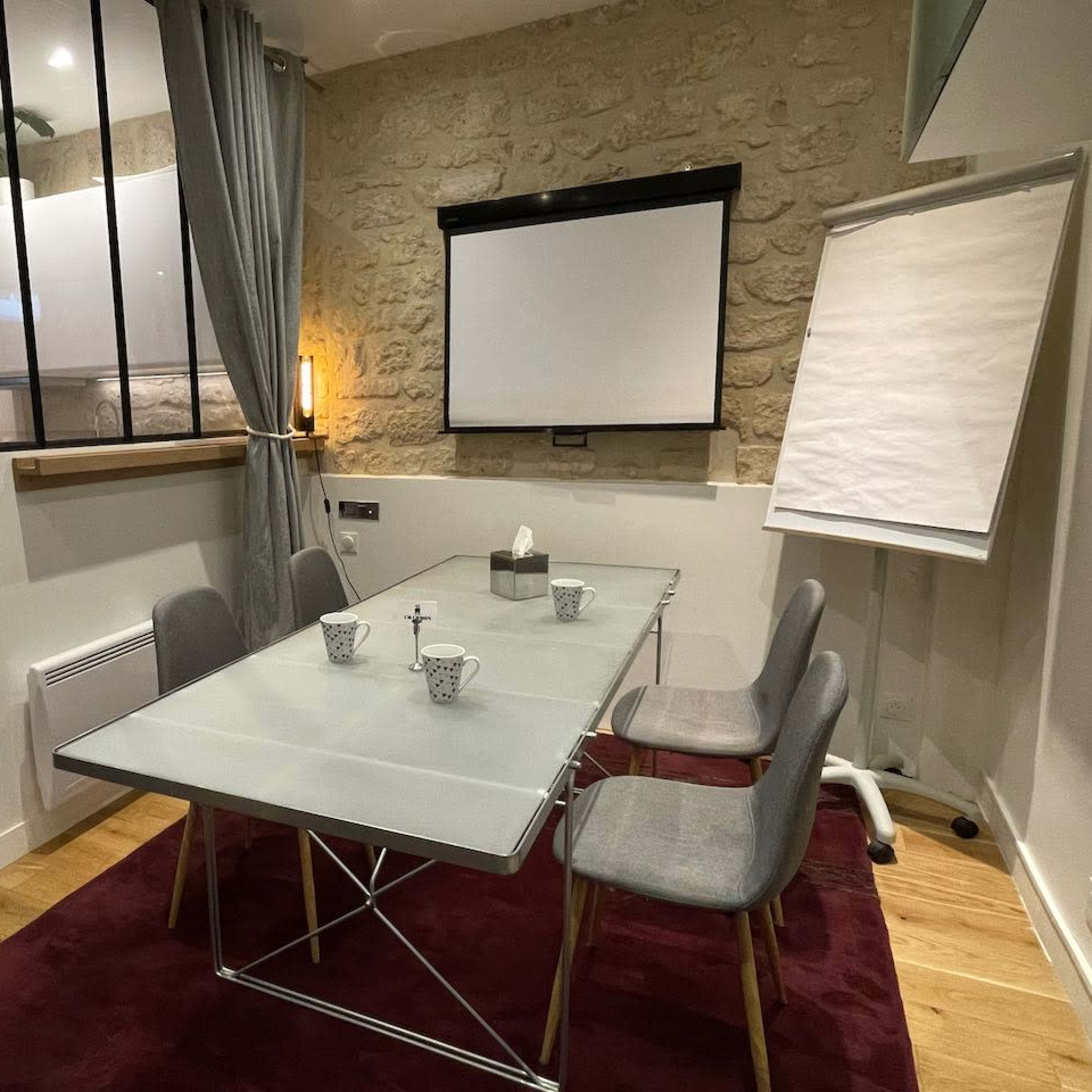 A glass-top conference table with four gray chairs is set up in a room with stone walls and a projector screen at one end.