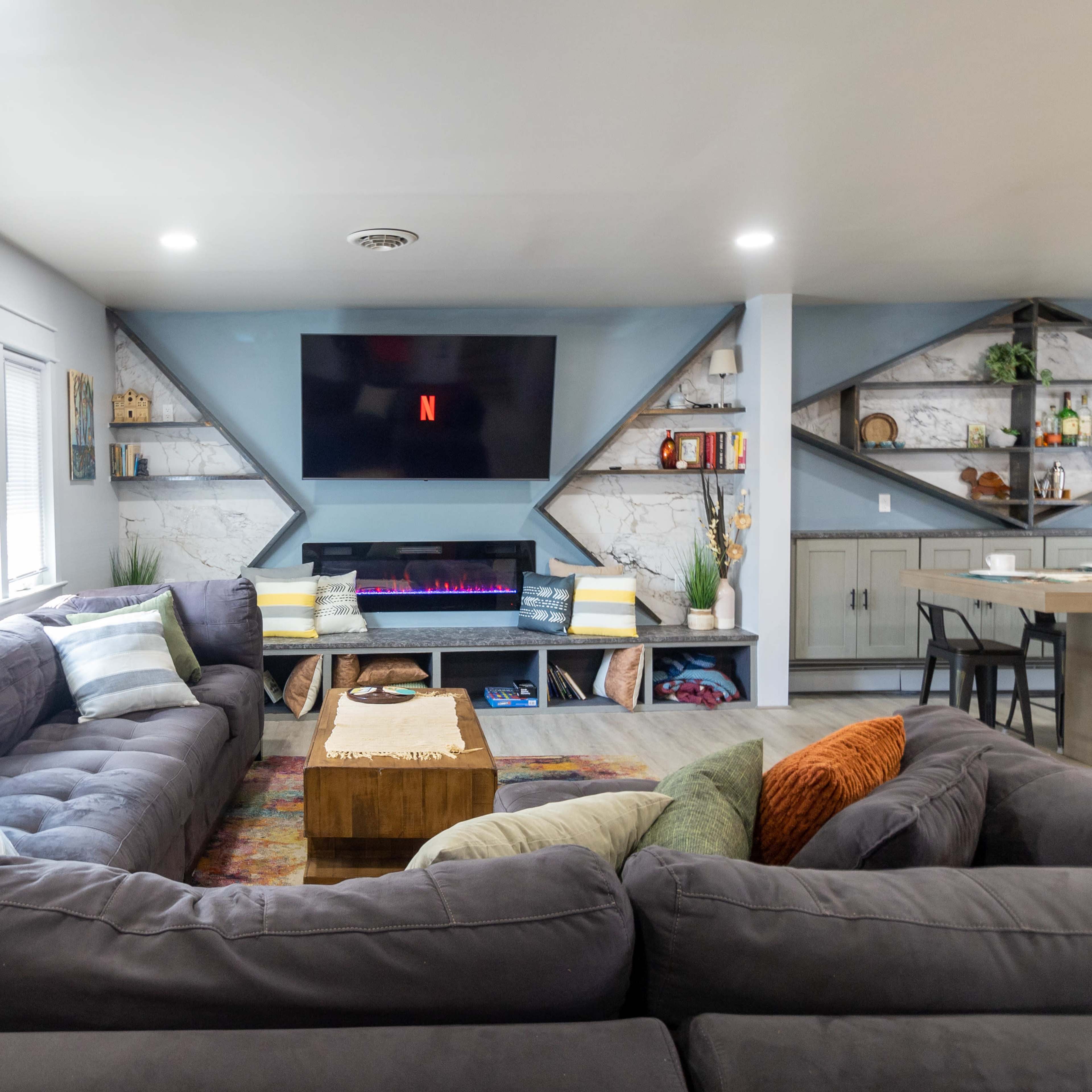 A modern living room with a large sectional sofa, a wall-mounted TV displaying the Netflix logo, and shelves filled with various decorative items and bottles.